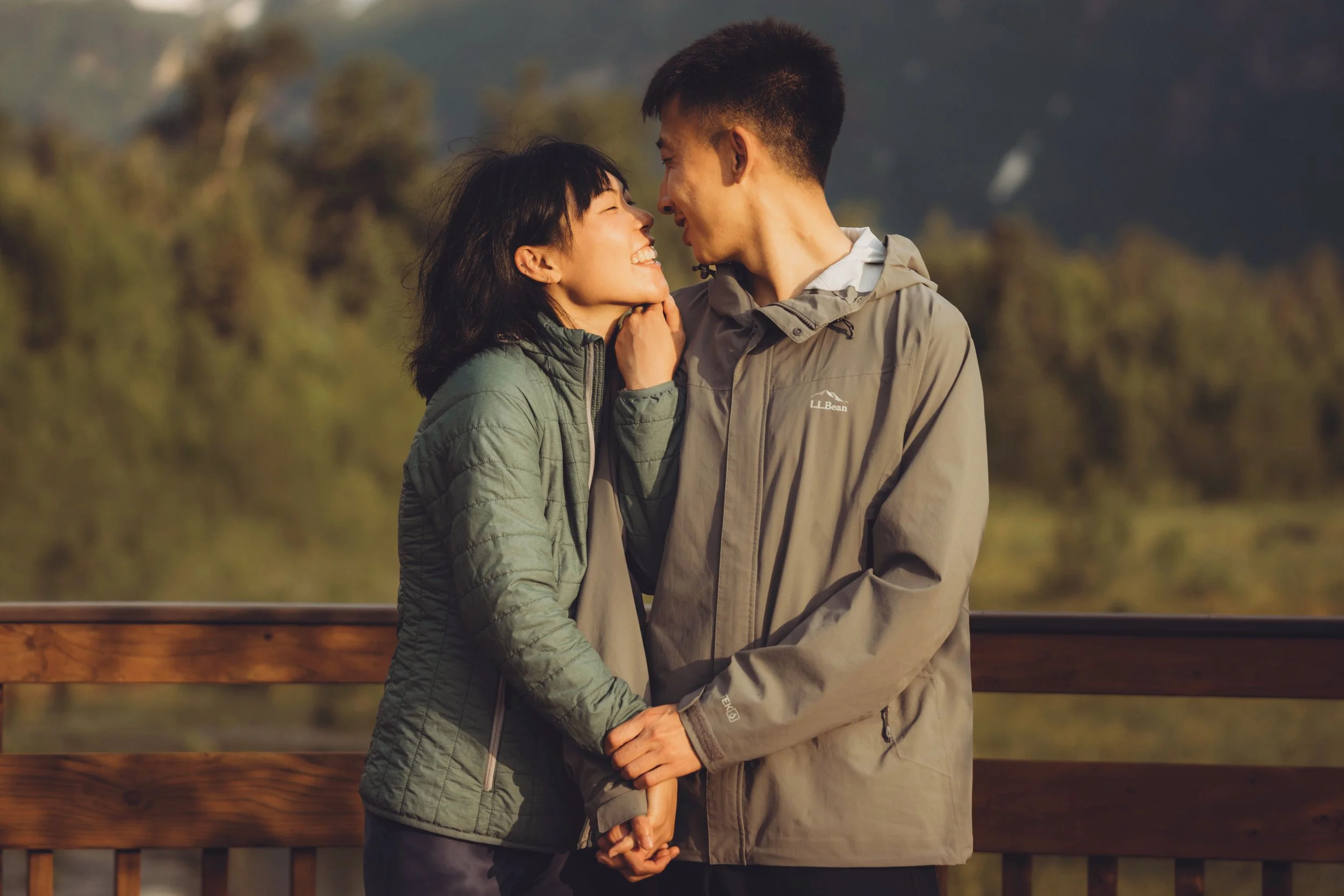 A couple smiling and gazing at each other, holding hands on a wooden railing outdoors with trees and mountains in the background.