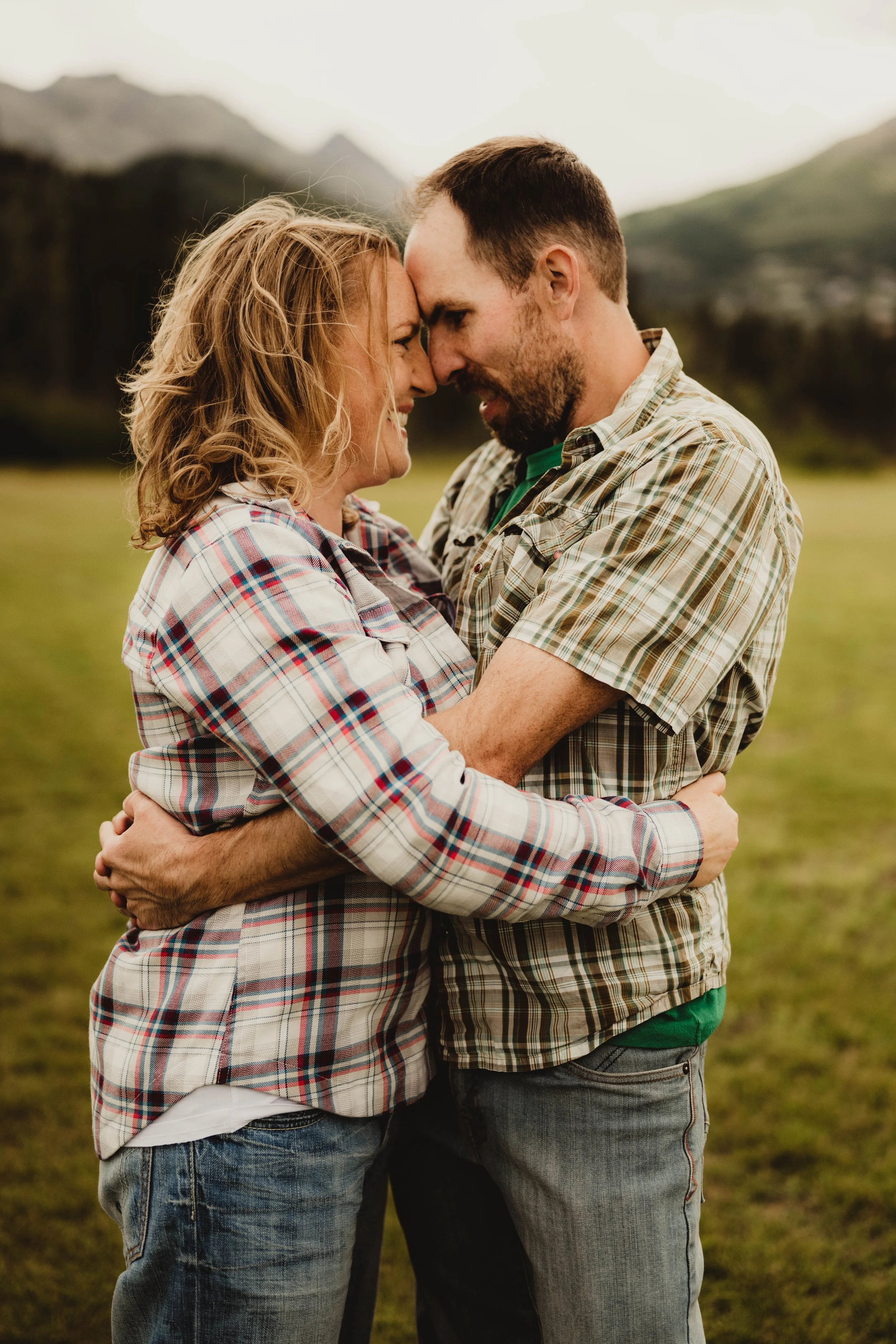 A couple hugging outdoors with their foreheads touching, smiling, and embracing each other, with mountains in the background on a cloudy day.
