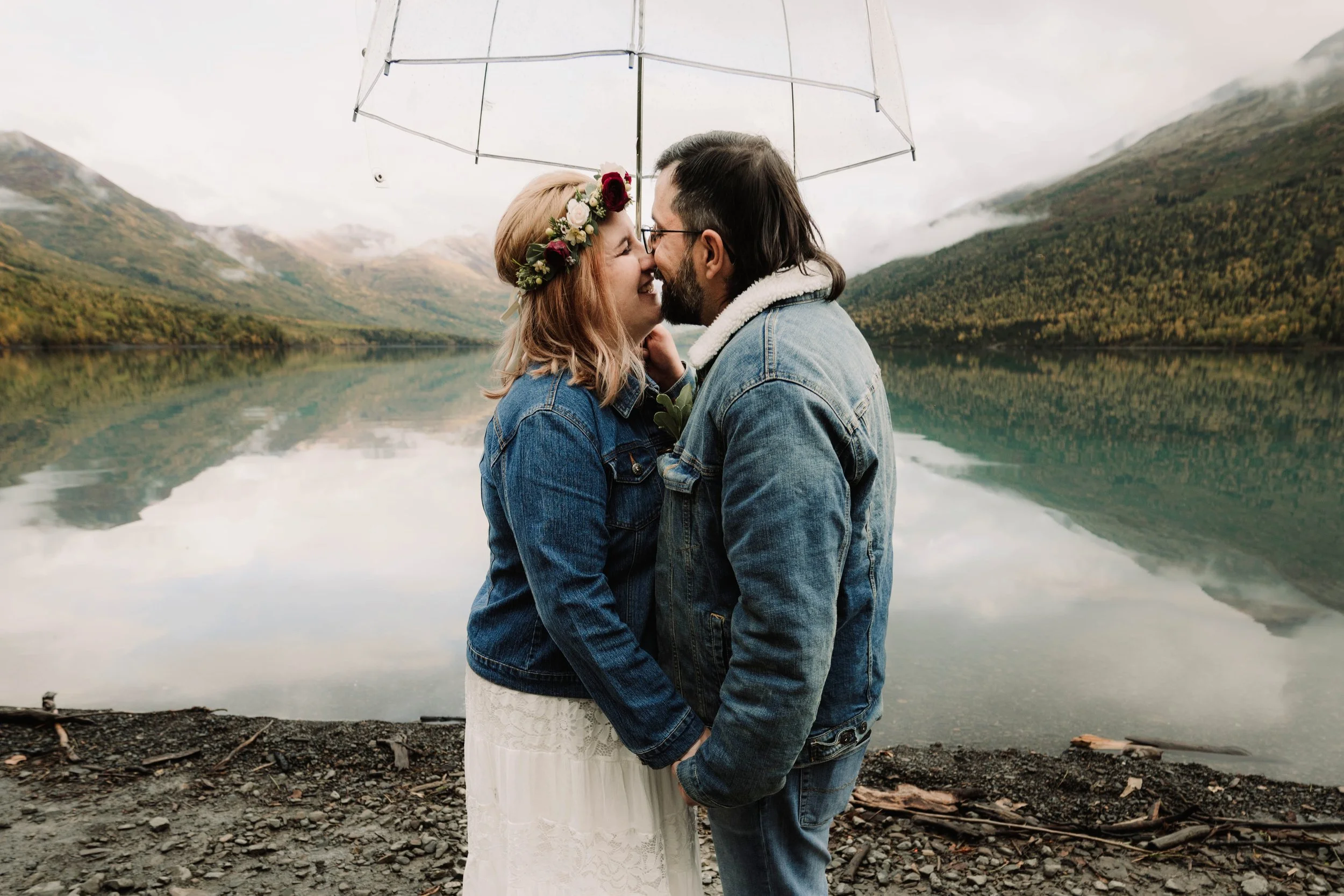 A couple standing close together with noses touching, sharing an umbrella by a lake surrounded by mountains and overcast skies.