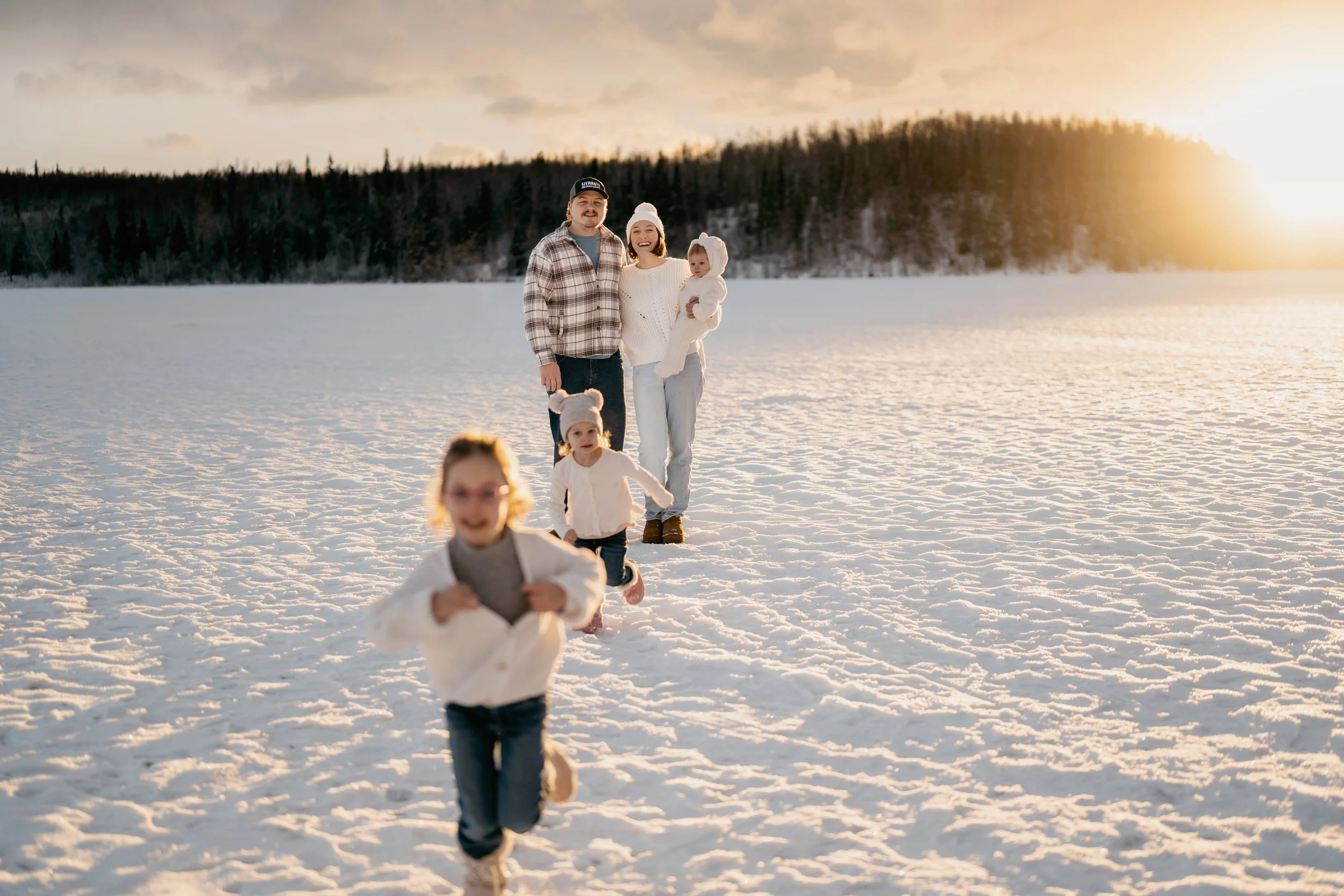 A family of five, including two children and two adults, enjoying a walk on a snowy field during sunset, with trees in the background.