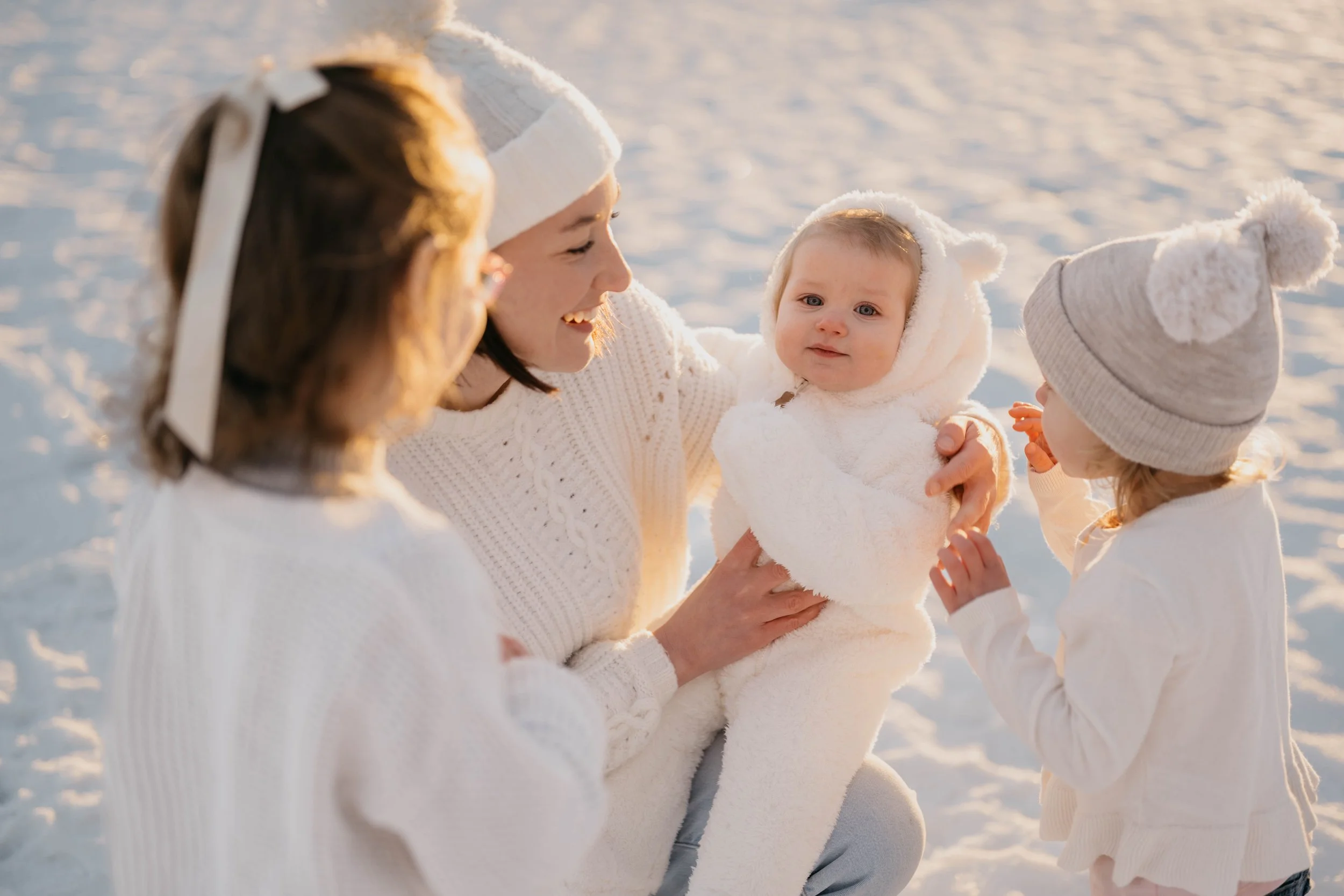 A woman holding a baby, surrounded by three children, all dressed in white winter clothing, in a snowy outdoor setting during sunset.