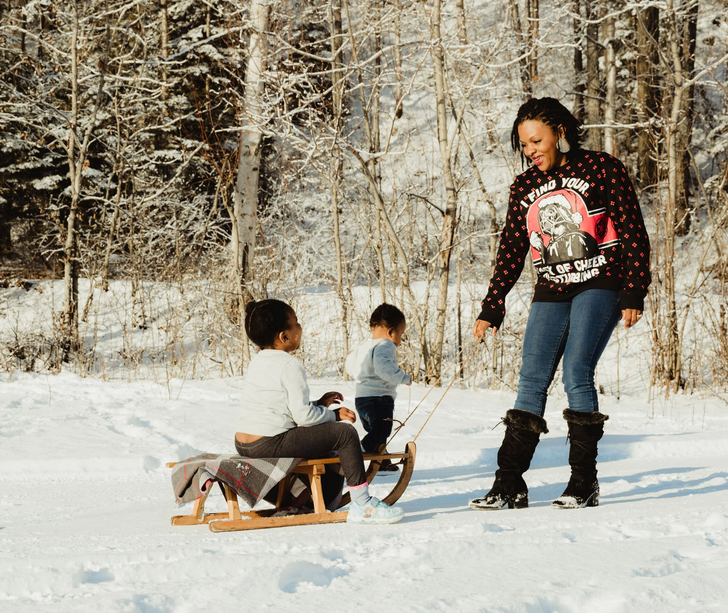 A woman in black winter boots and a black sweater with a graphic and text is pulling a sled with two children in a snowy forested area.