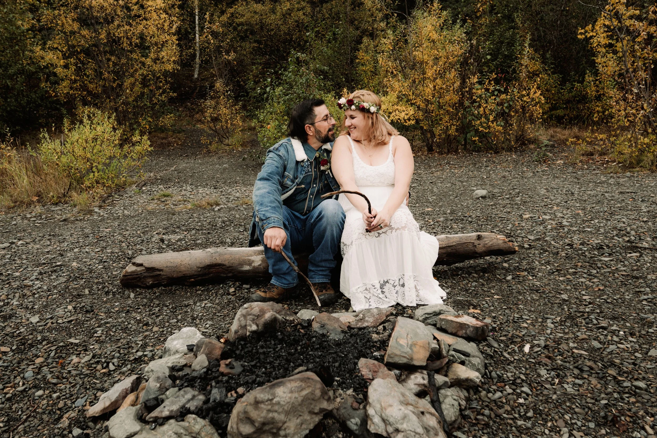 A couple sitting on a log by a campfire in a forest, smiling at each other. The woman is wearing a white lace dress and a flower crown, and the man is wearing a denim jacket and jeans.
