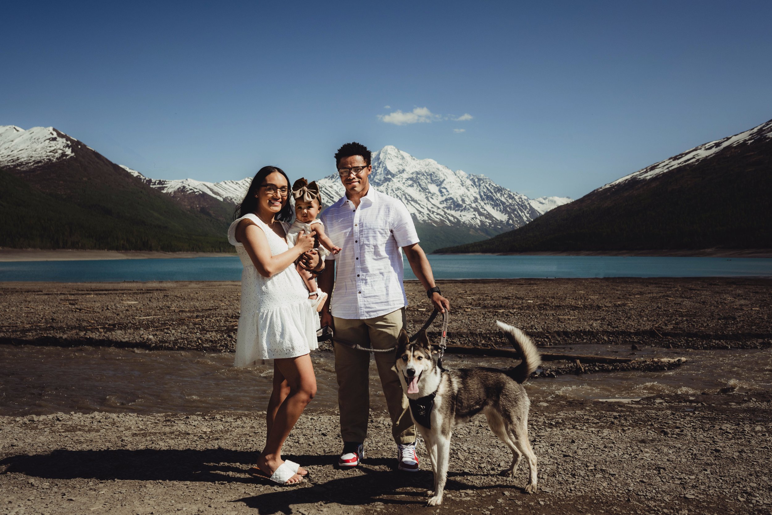 A family of three with a husky dog standing by a lake surrounded by snow-capped mountains and green hills under a clear blue sky.