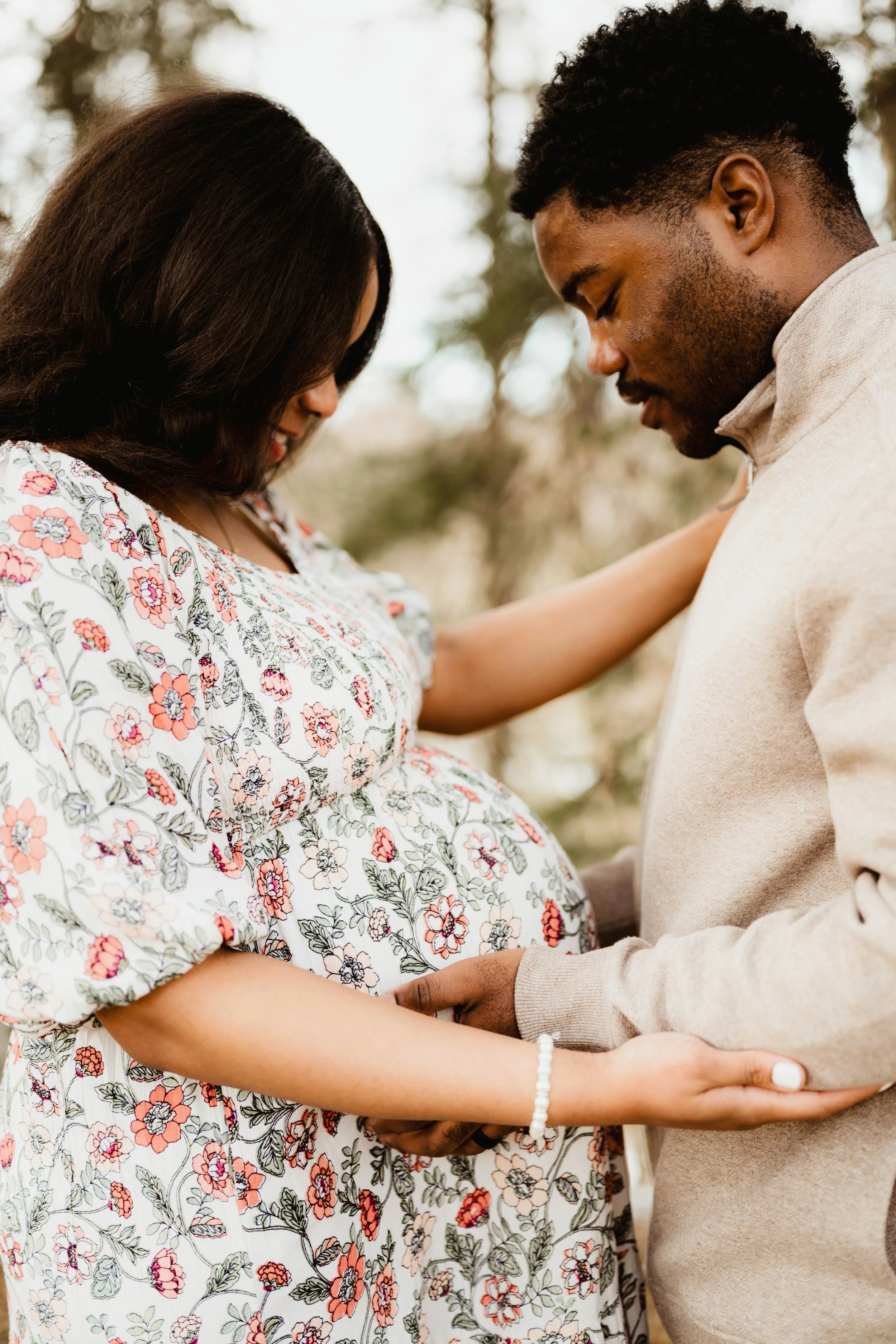 A pregnant woman in a floral dress and a man in a beige sweater hold hands and share a tender moment outdoors, with their foreheads touching and eyes closed.