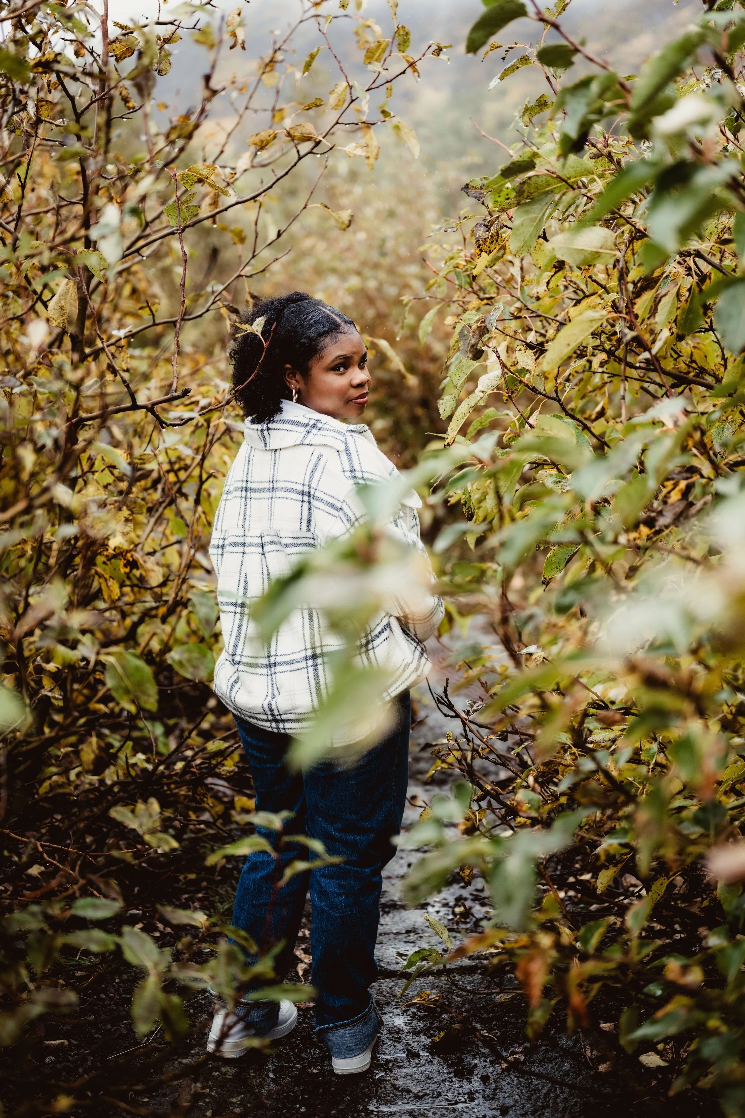 A woman standing on a muddy trail surrounded by dense, yellowing foliage in an outdoor setting.