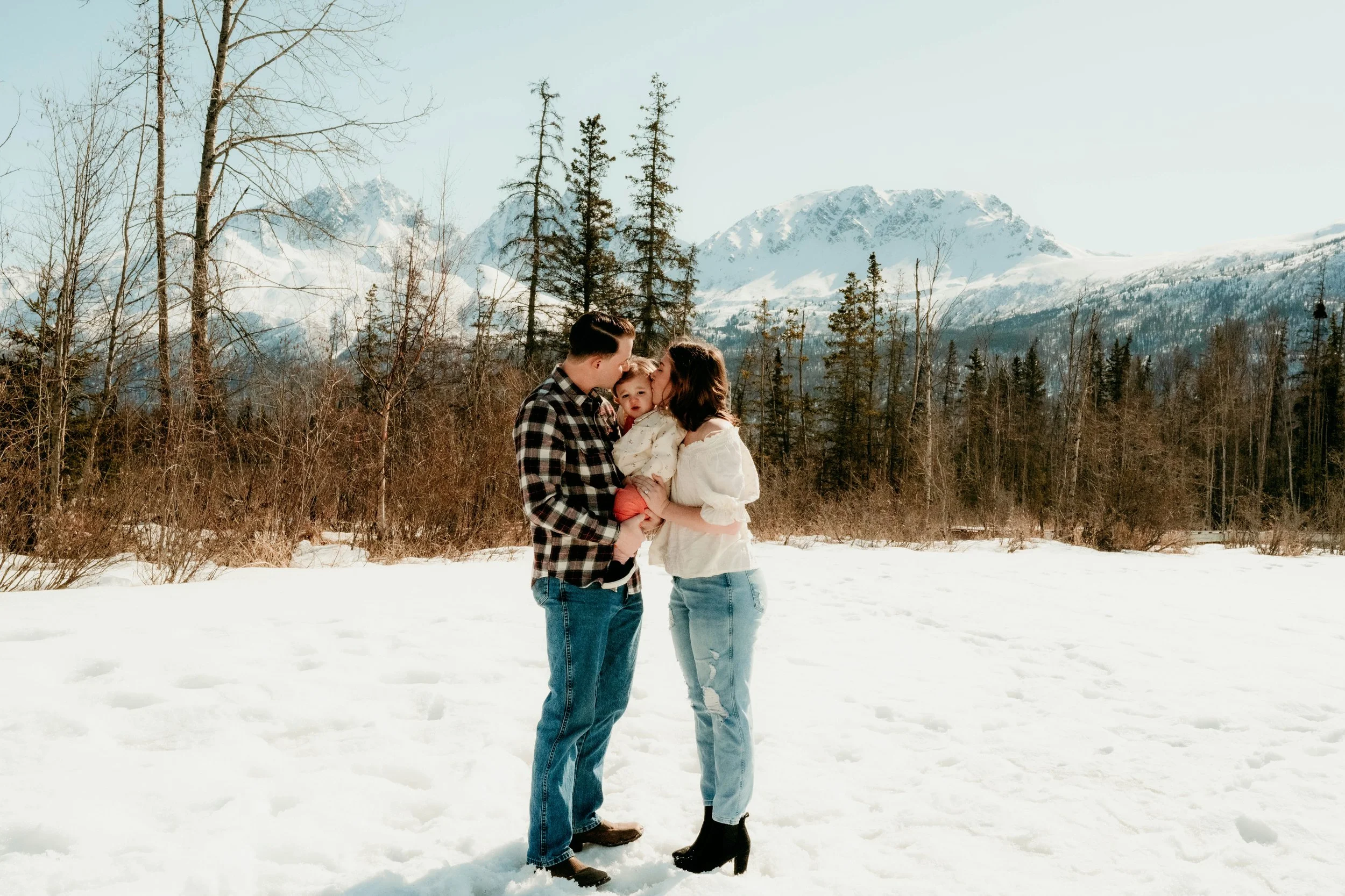 A family of three standing in a snowy landscape with mountains and trees in the background. The man and woman are holding their young daughter, who appears to be reaching toward them.