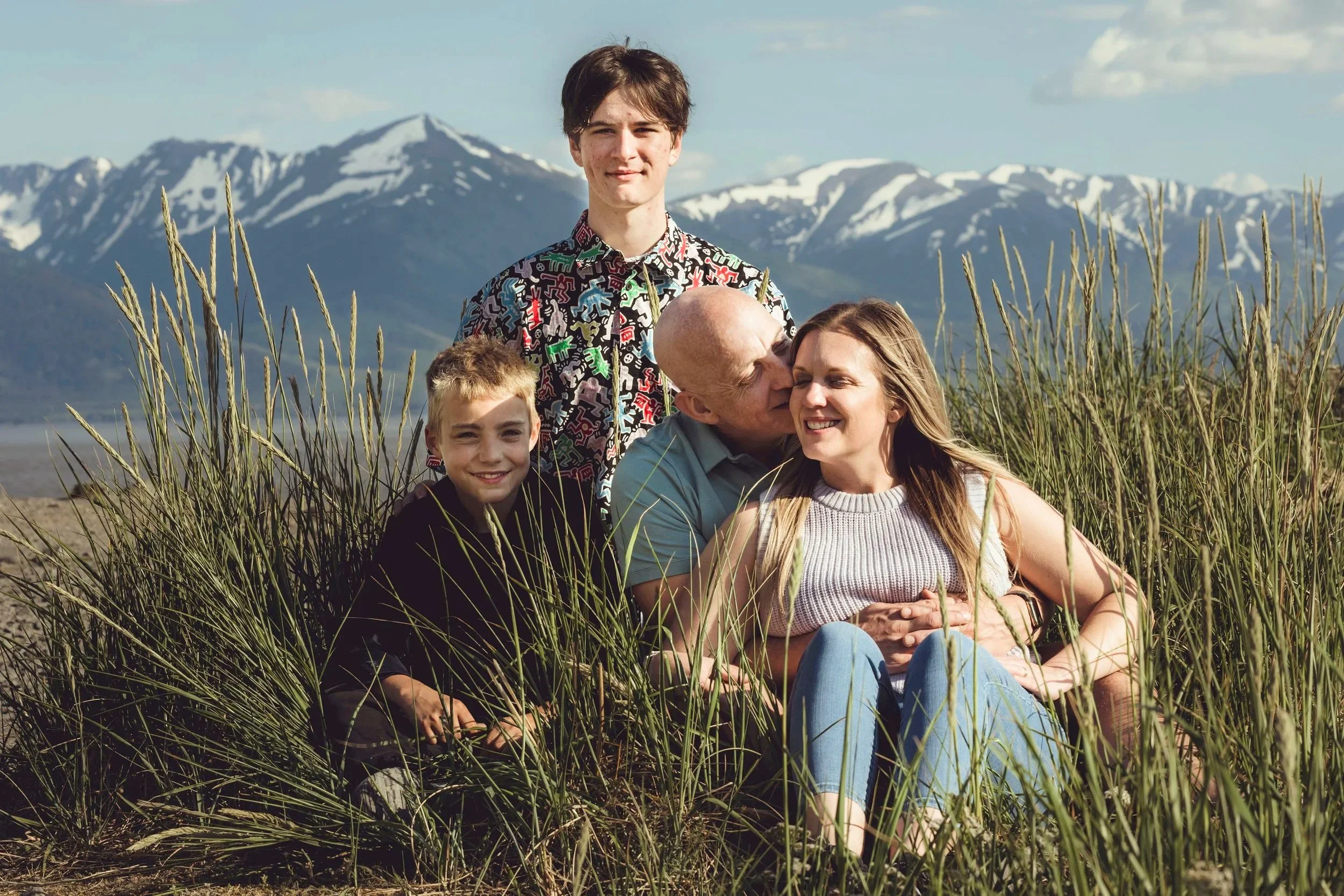 A family of four sitting in tall grass with snow-capped mountains in the background, smiling and enjoying time together outdoors.