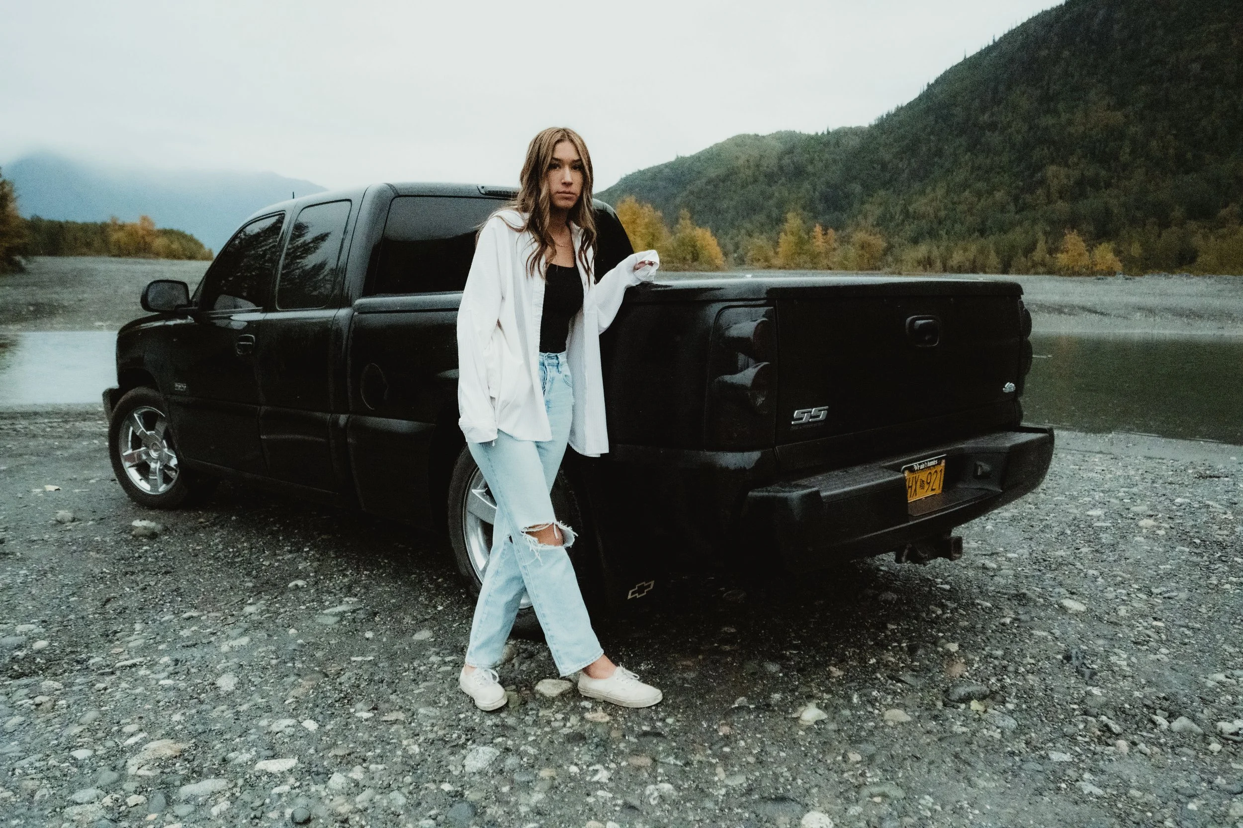 A woman leaning against the bed of a black pickup truck near a river, with mountains and trees in the background, dressed in casual clothing.