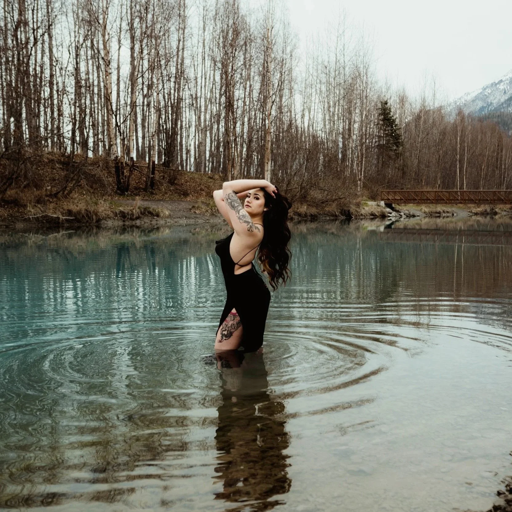 A woman with dark hair and tattoos stands in a body of water, wearing a black dress, with a forest and mountains in the background.