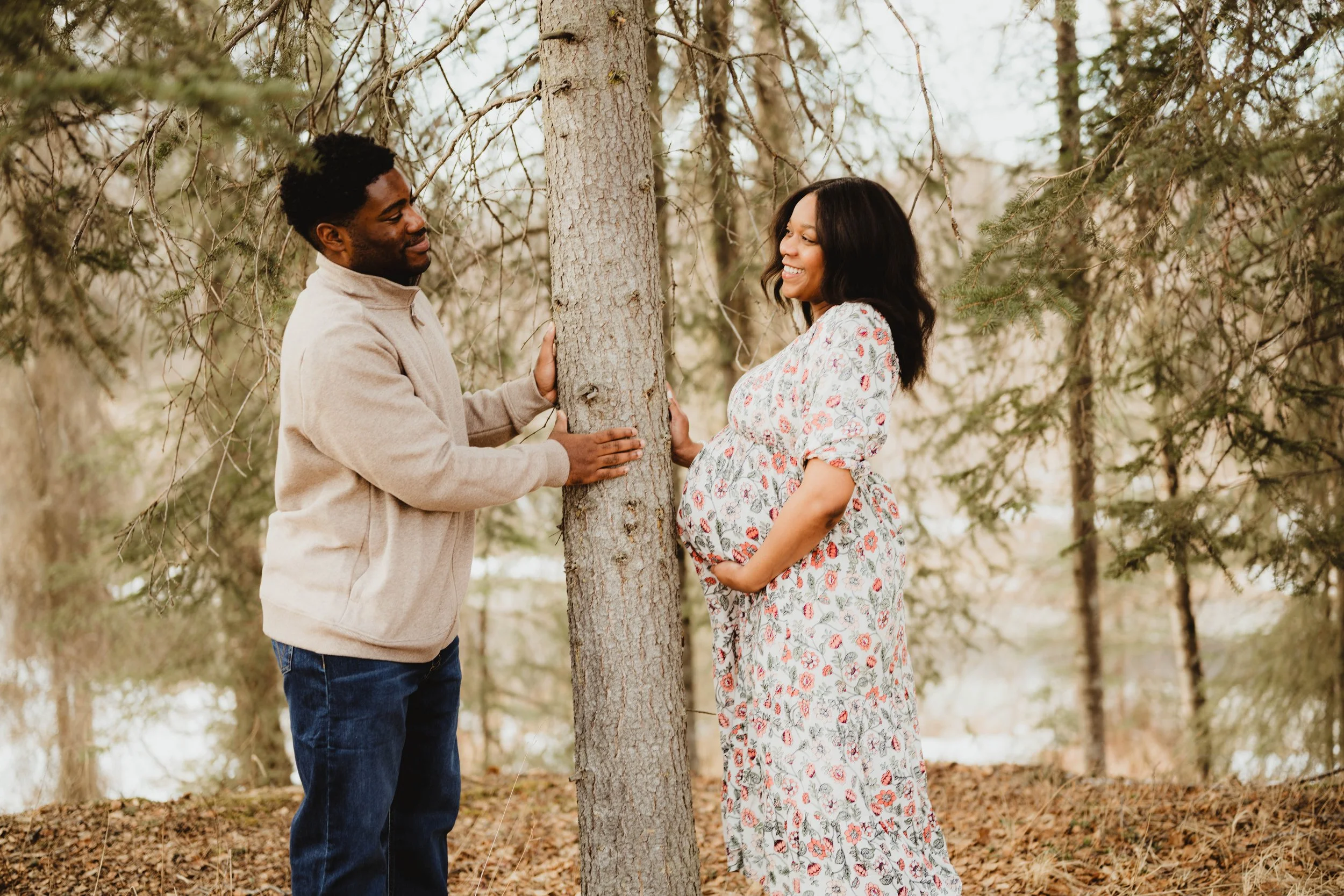 A man and pregnant woman standing behind a tree, smiling at each other in a forested area.