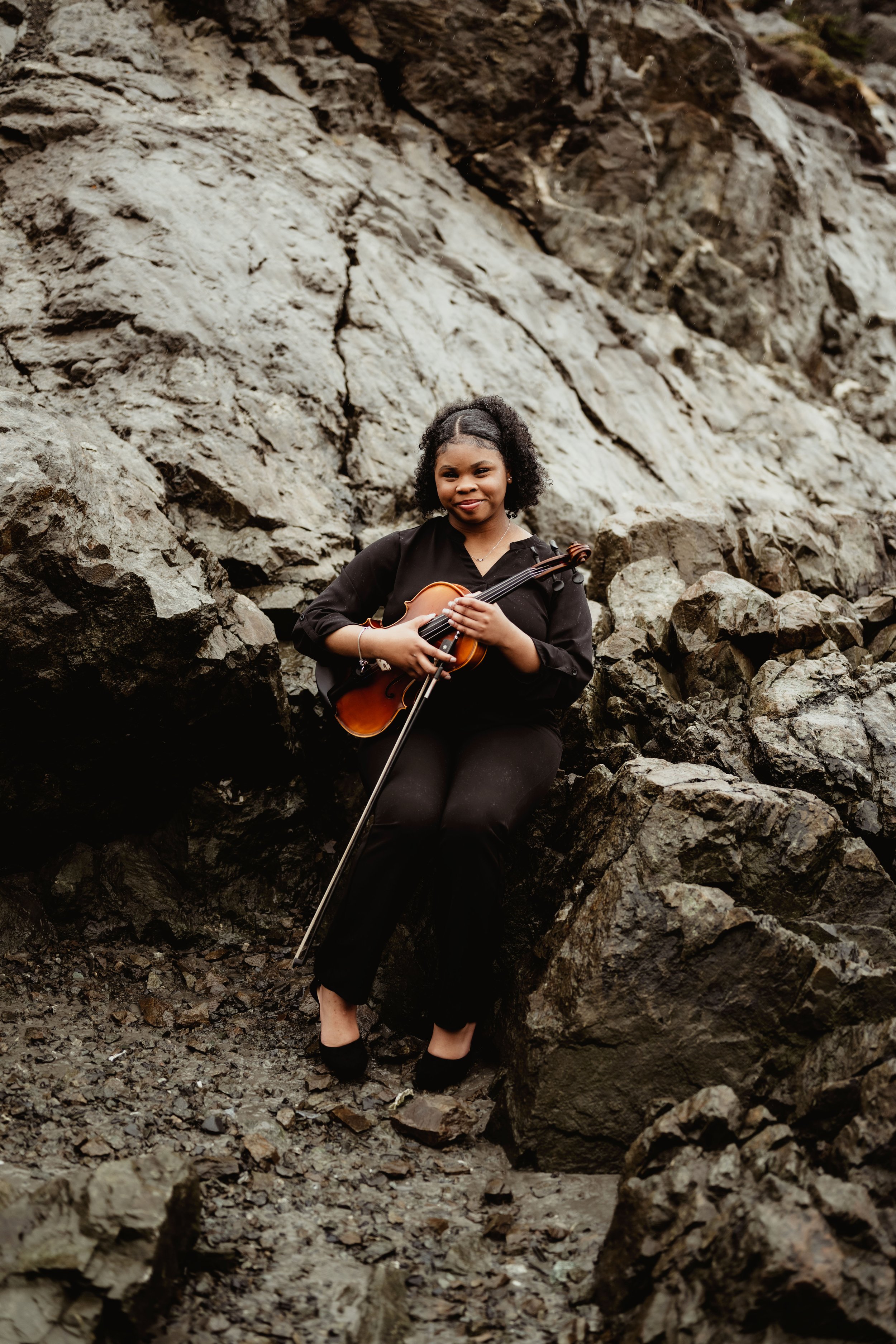 A woman in black clothing sitting on rocks, holding a violin and bow, with a rocky background.
