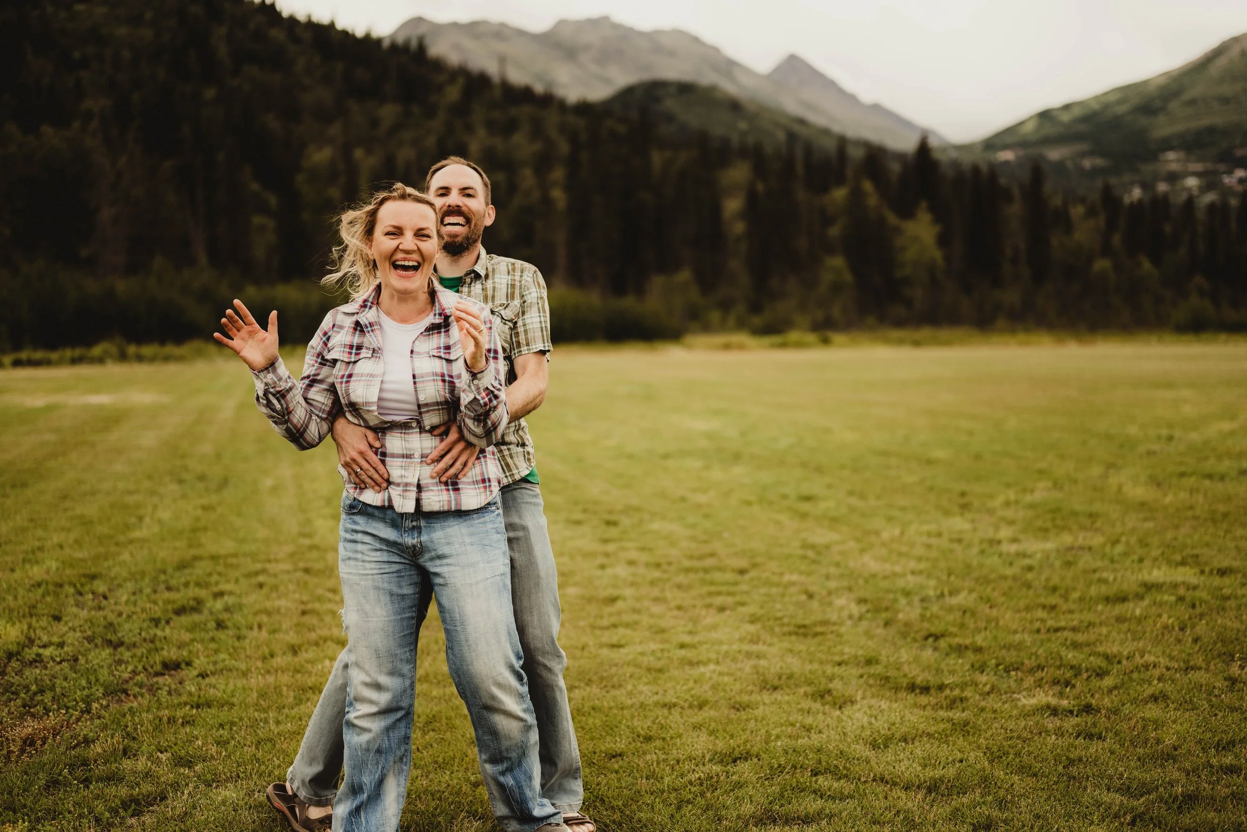 A happy couple laughing in an open grassy field with mountains and trees in the background.