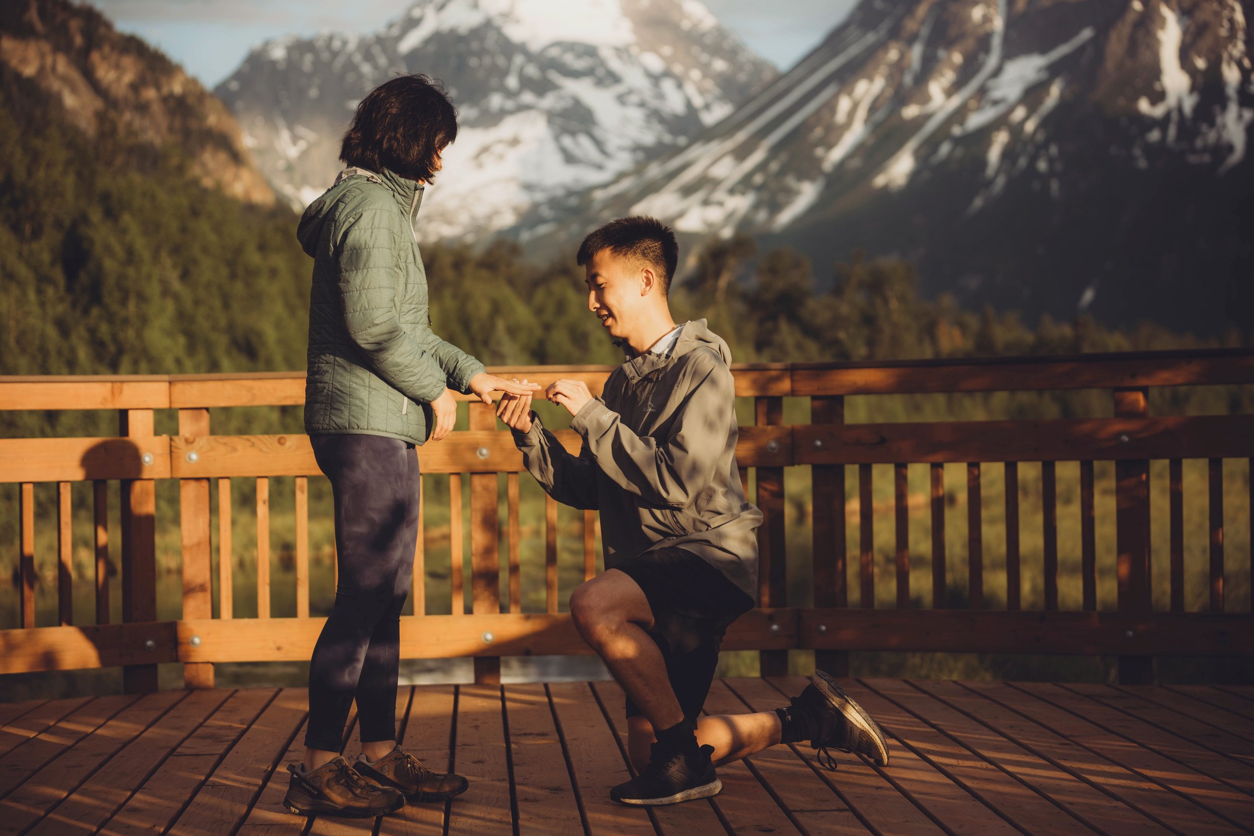 A man proposing marriage to a woman on a wooden deck with mountains in the background during sunset.