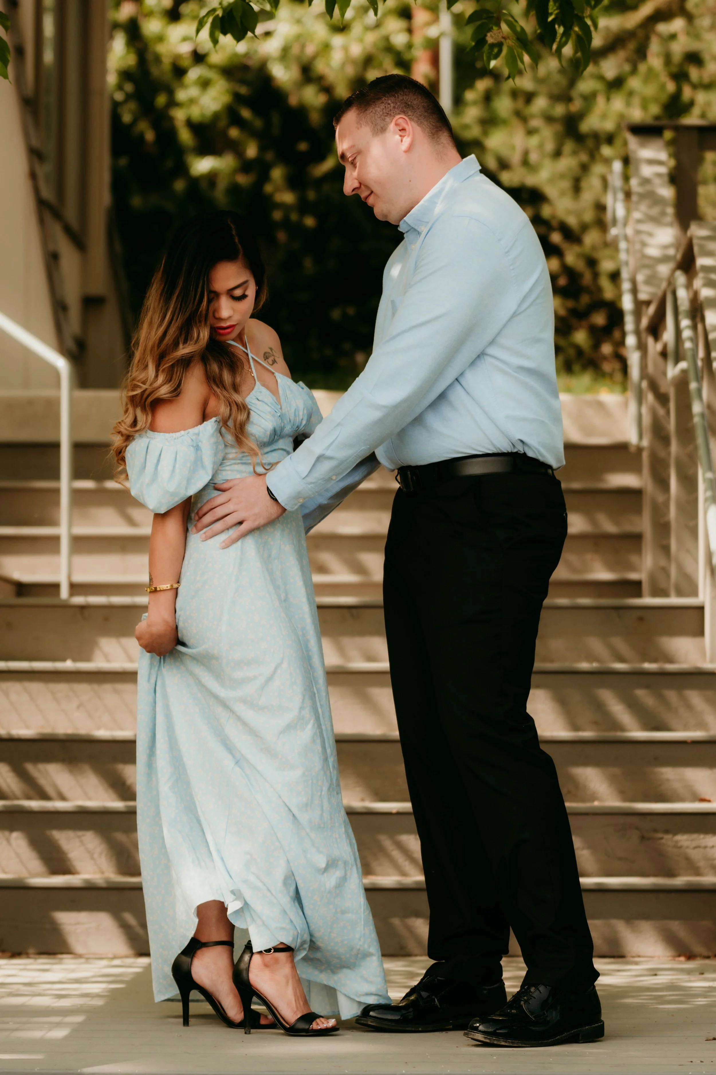 A man and woman standing on outdoor steps, the man gently holds the woman's waist, both looking down with a tender expression. The woman is wearing a light blue off-shoulder dress and black high heels, while the man is dressed in a light blue shirt and black pants.