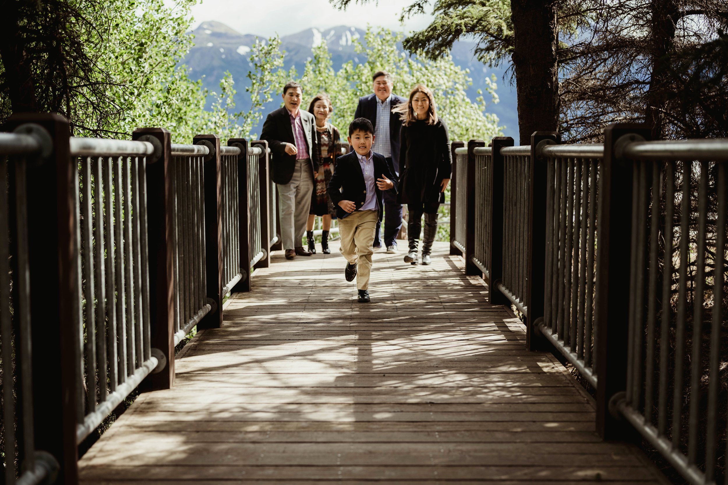 A young boy in a blazer running across a wooden bridge with five adults following behind, set against a backdrop of green trees and distant mountains.