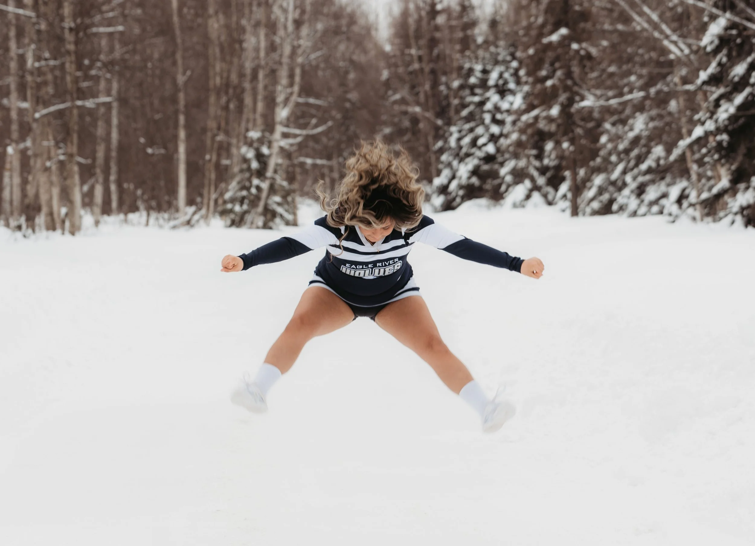 A person wearing sports apparel and white socks standing in snow with legs apart and arms extended, in a snowy forest landscape.