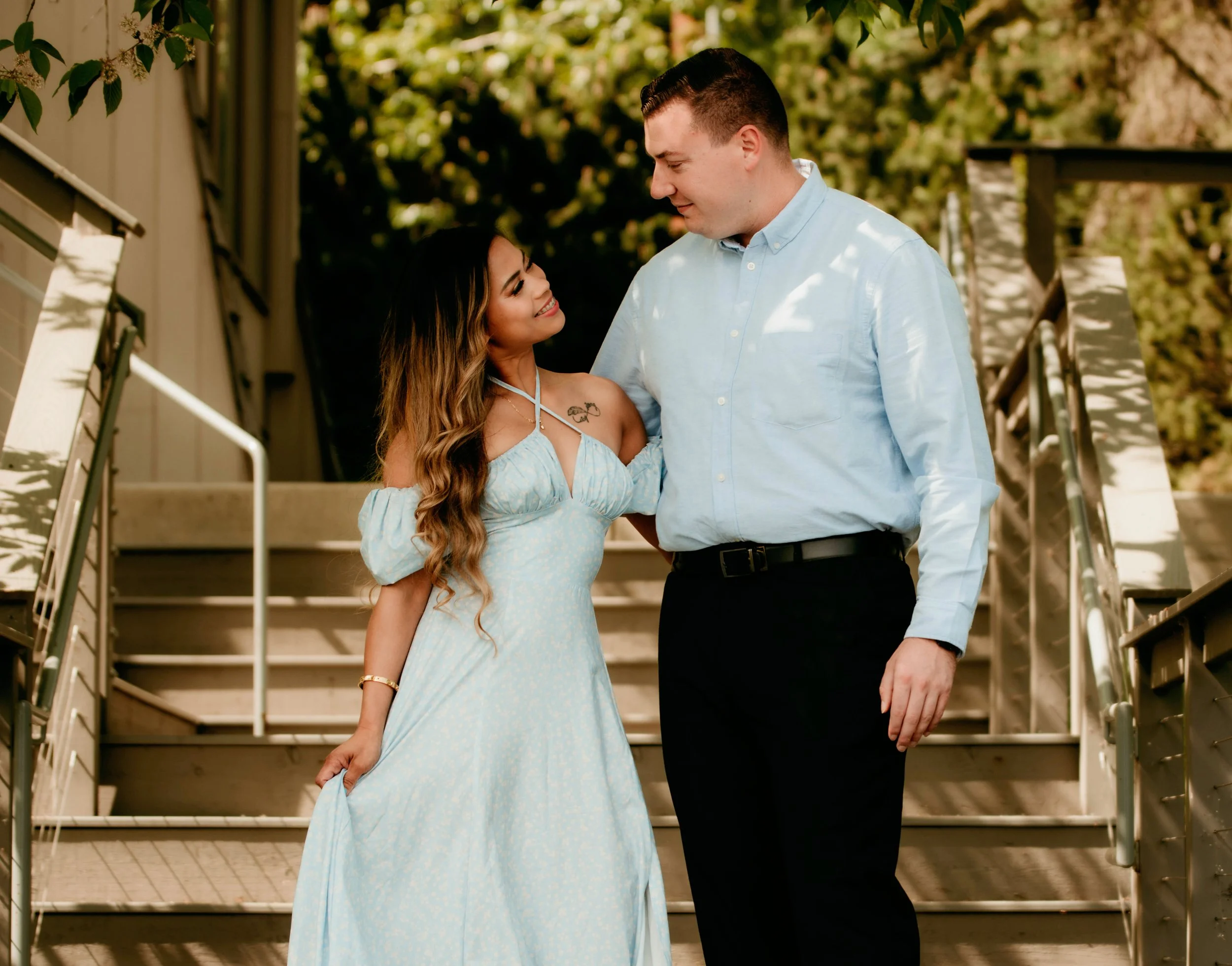 A woman in a light blue dress looking at a man in a light blue shirt on an outdoor staircase, smiling, with trees in the background.
