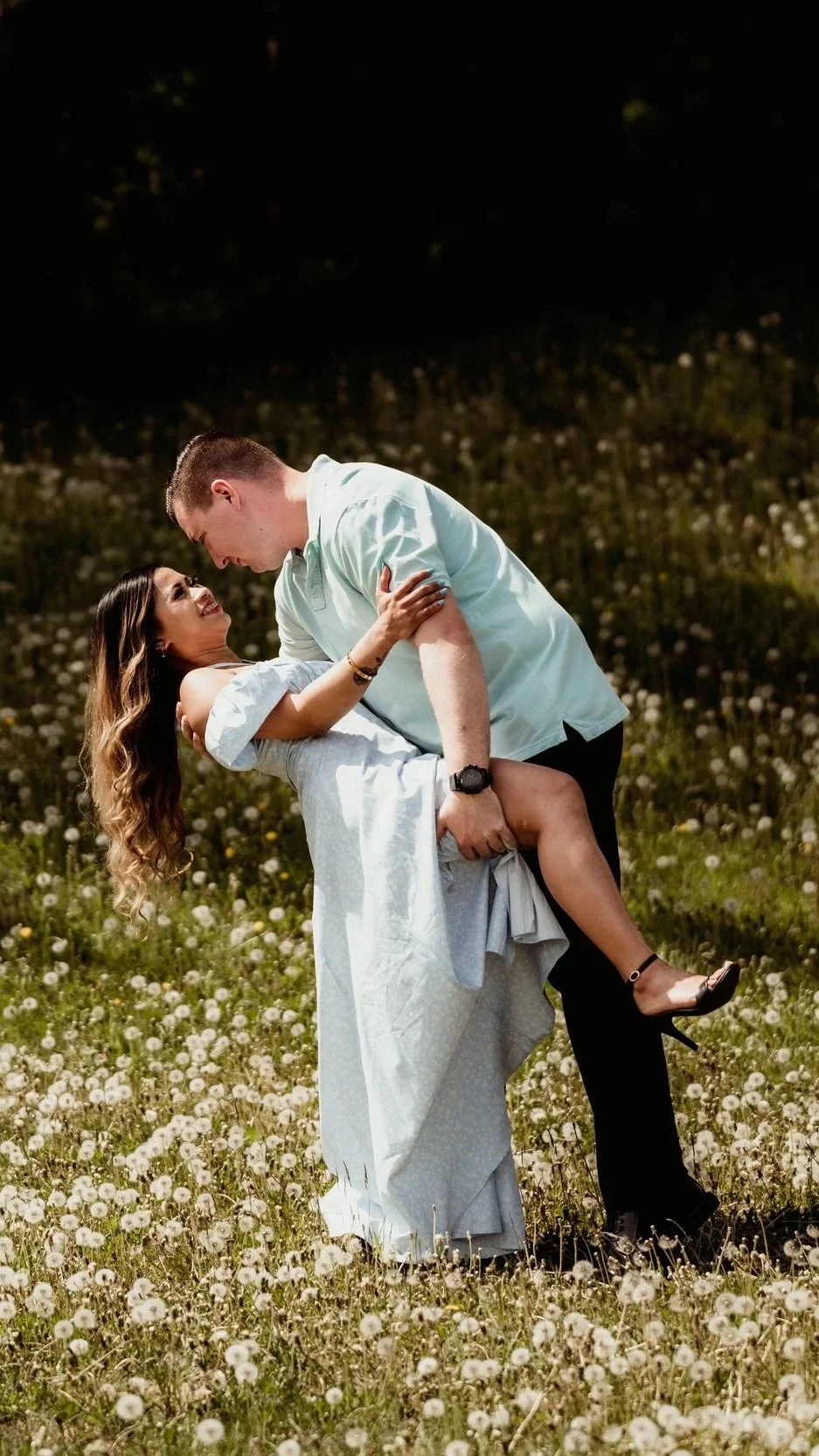 A man and woman dancing in a field of white flowers during sunset, with the man lifting the woman in his arms.