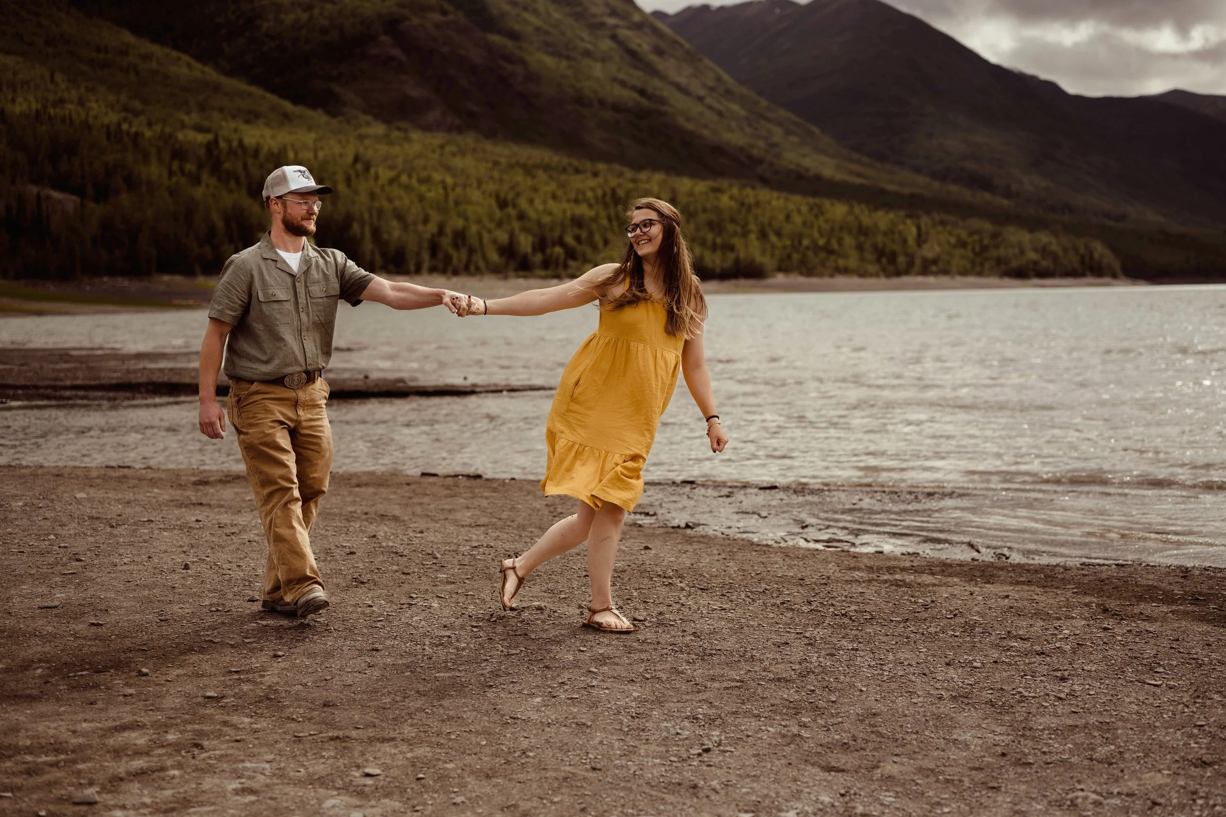 A young couple holding hands and dancing on a lakeshore with mountains and trees in the background.
