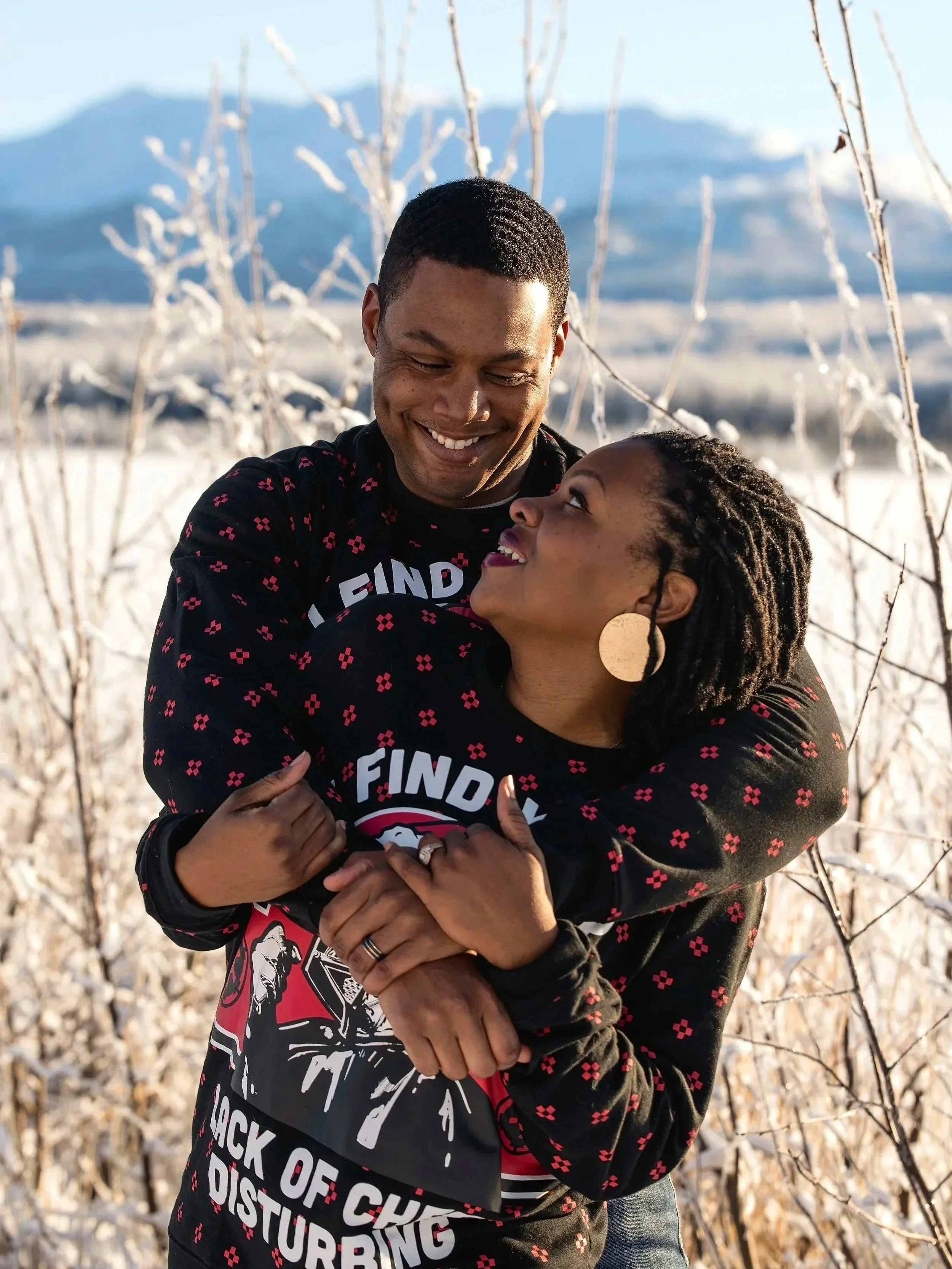 A happy couple embracing outdoors in a snowy landscape with mountains in the background. They are smiling and looking at each other.