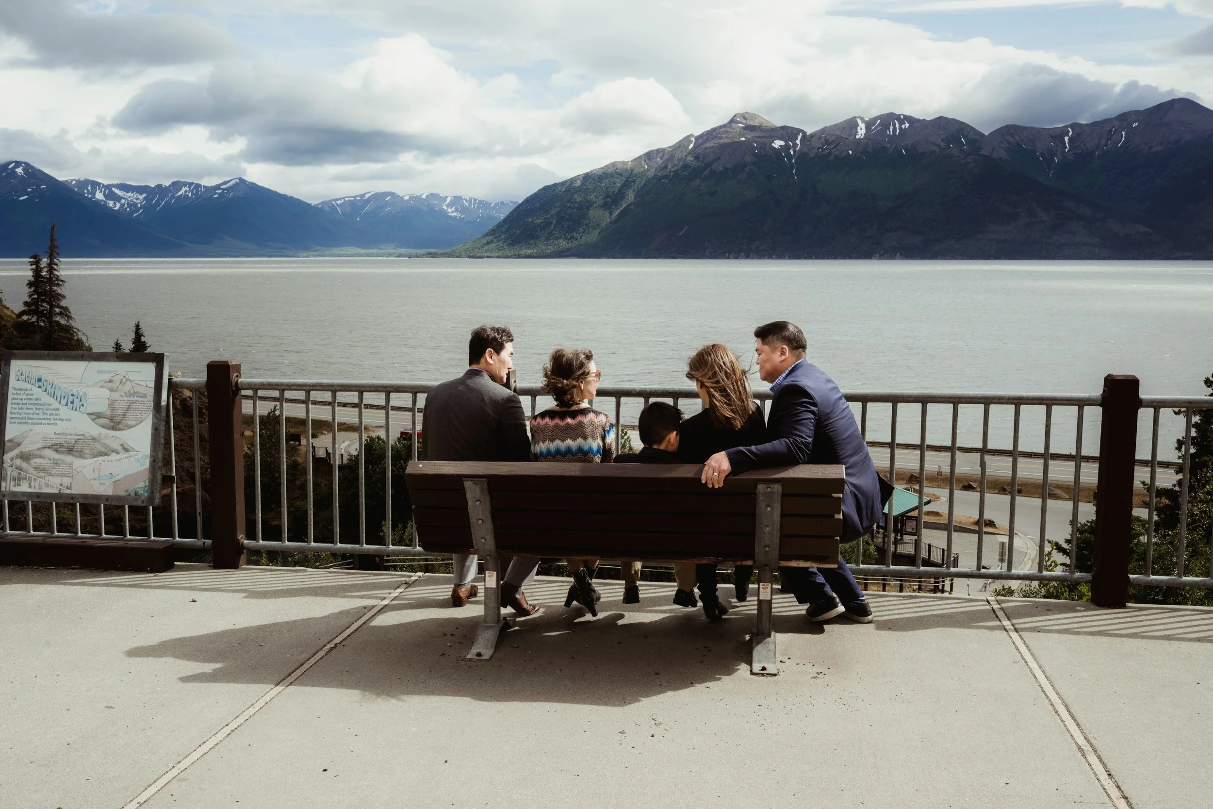 A group of six people sitting on a park bench, facing a large body of water with mountains in the background.