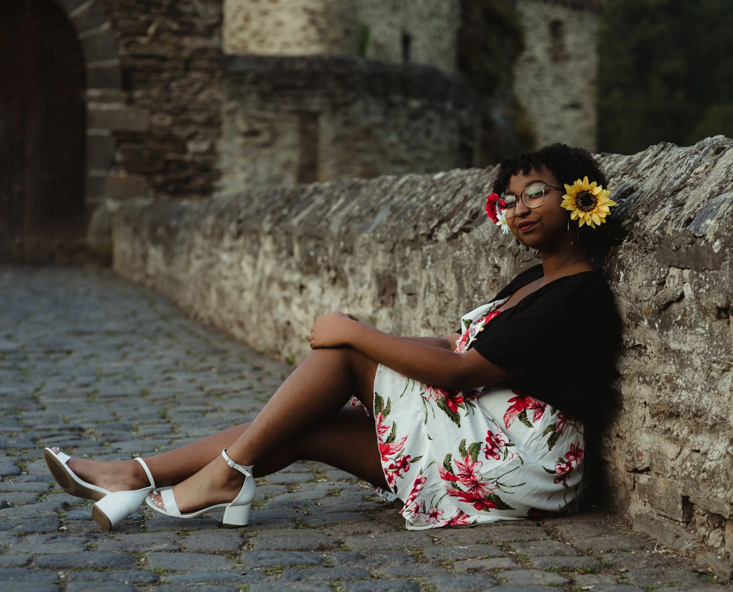 A woman with glasses and curly hair sitting on a cobblestone street, leaning against an old stone wall, wearing a floral dress, black cardigan, white high-heeled shoes, and flowers in her hair, including a sunflower and red flower.
