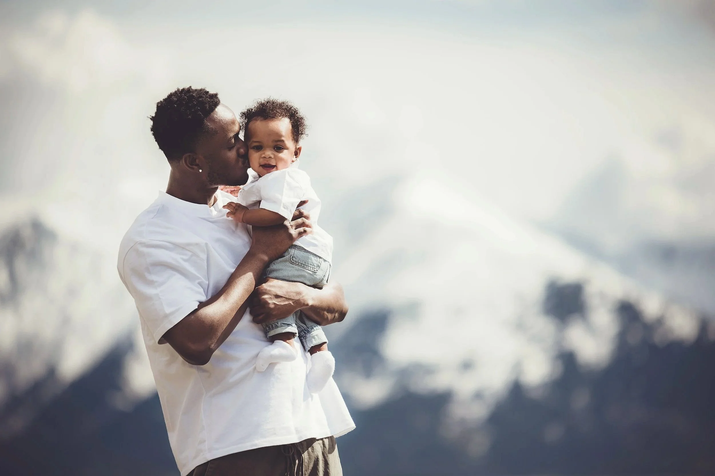 A man holds a baby and kisses the baby's cheek outdoors with mountains and cloudy sky in the background.