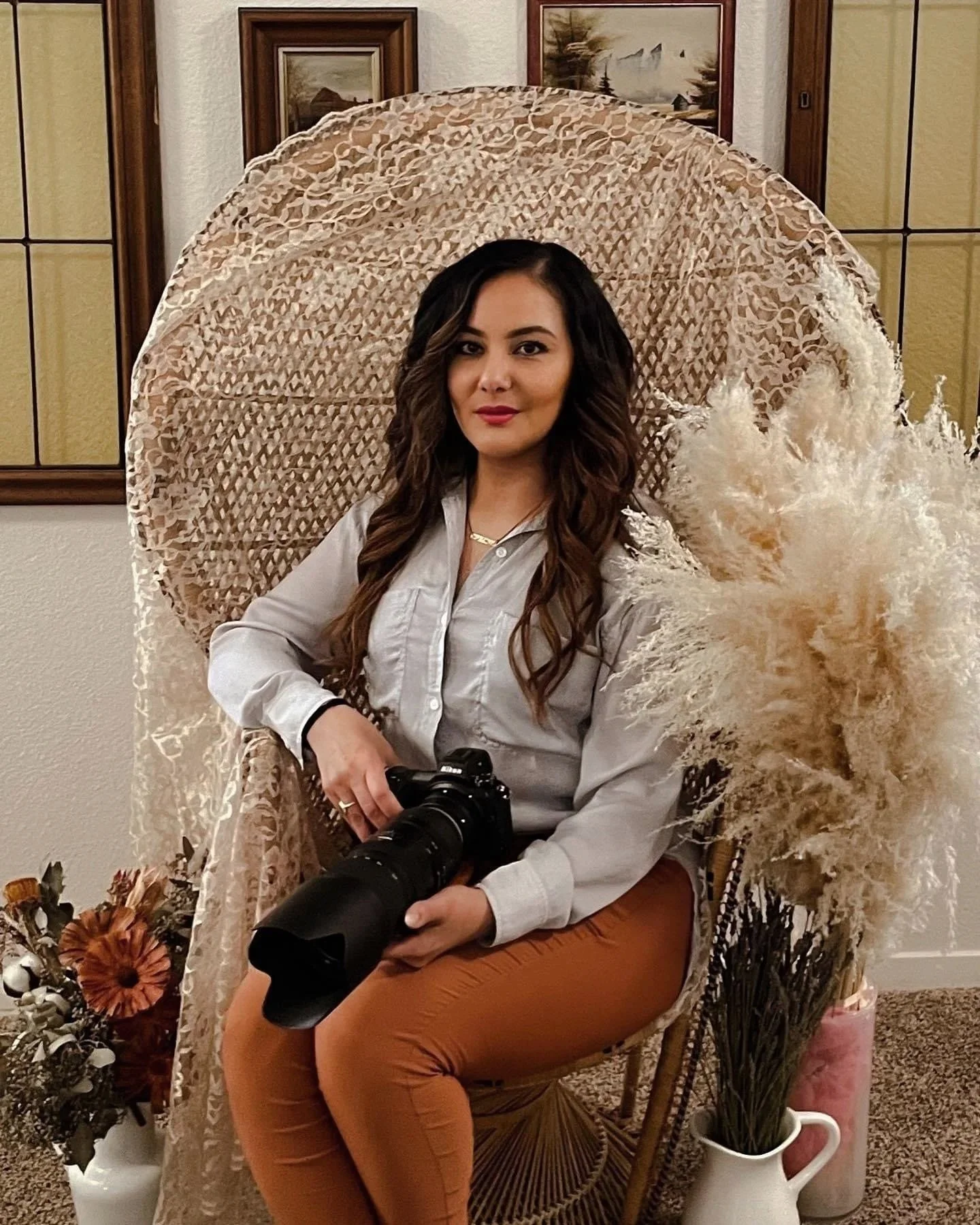 A woman with long wavy brown hair sitting on a wicker chair, holding a camera with a large lens, in a room with framed artwork and dried flowers.