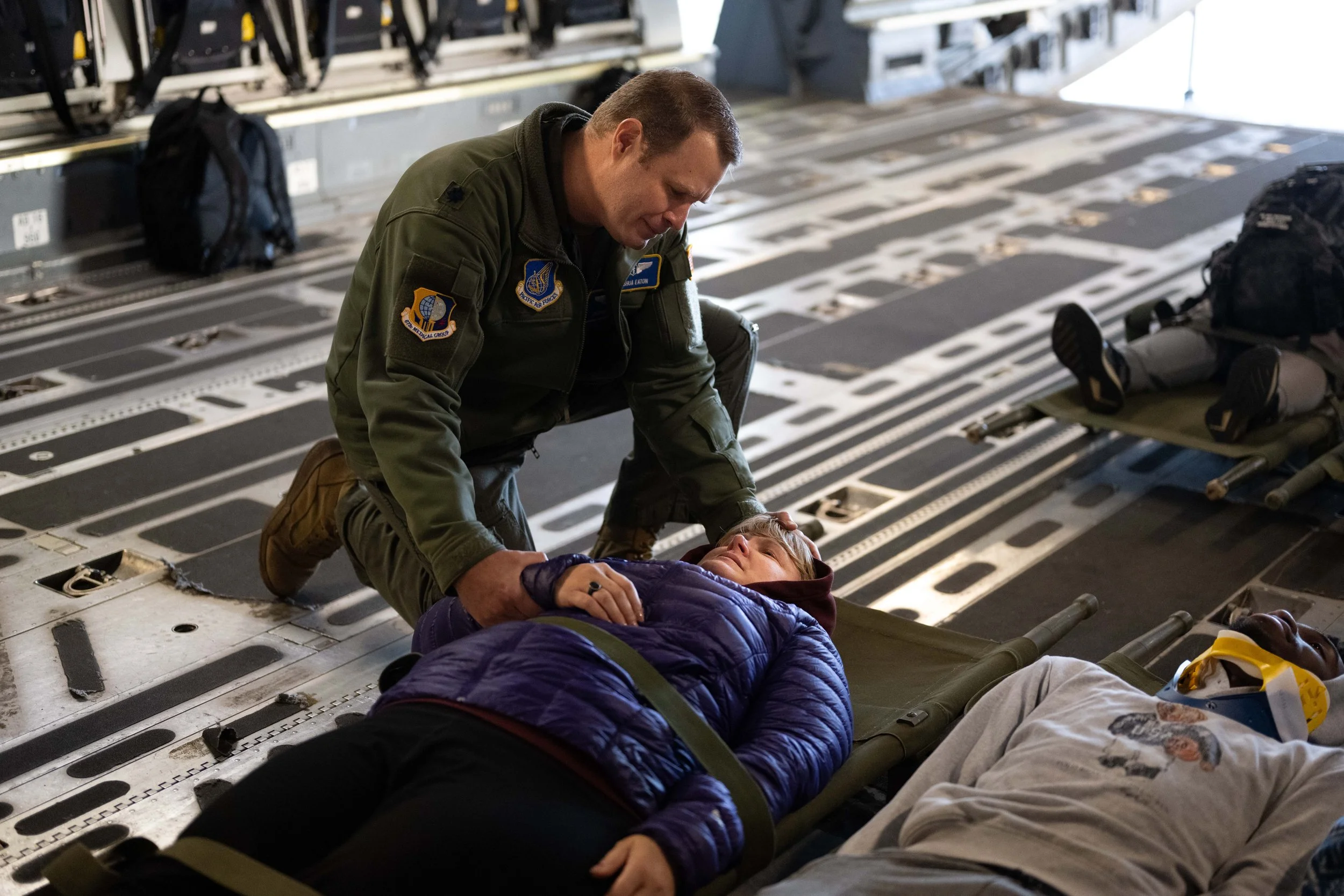 A man in military attire attending to a woman lying on a stretcher inside an aircraft, another person is lying nearby, suggesting a rescue or medical emergency scene.