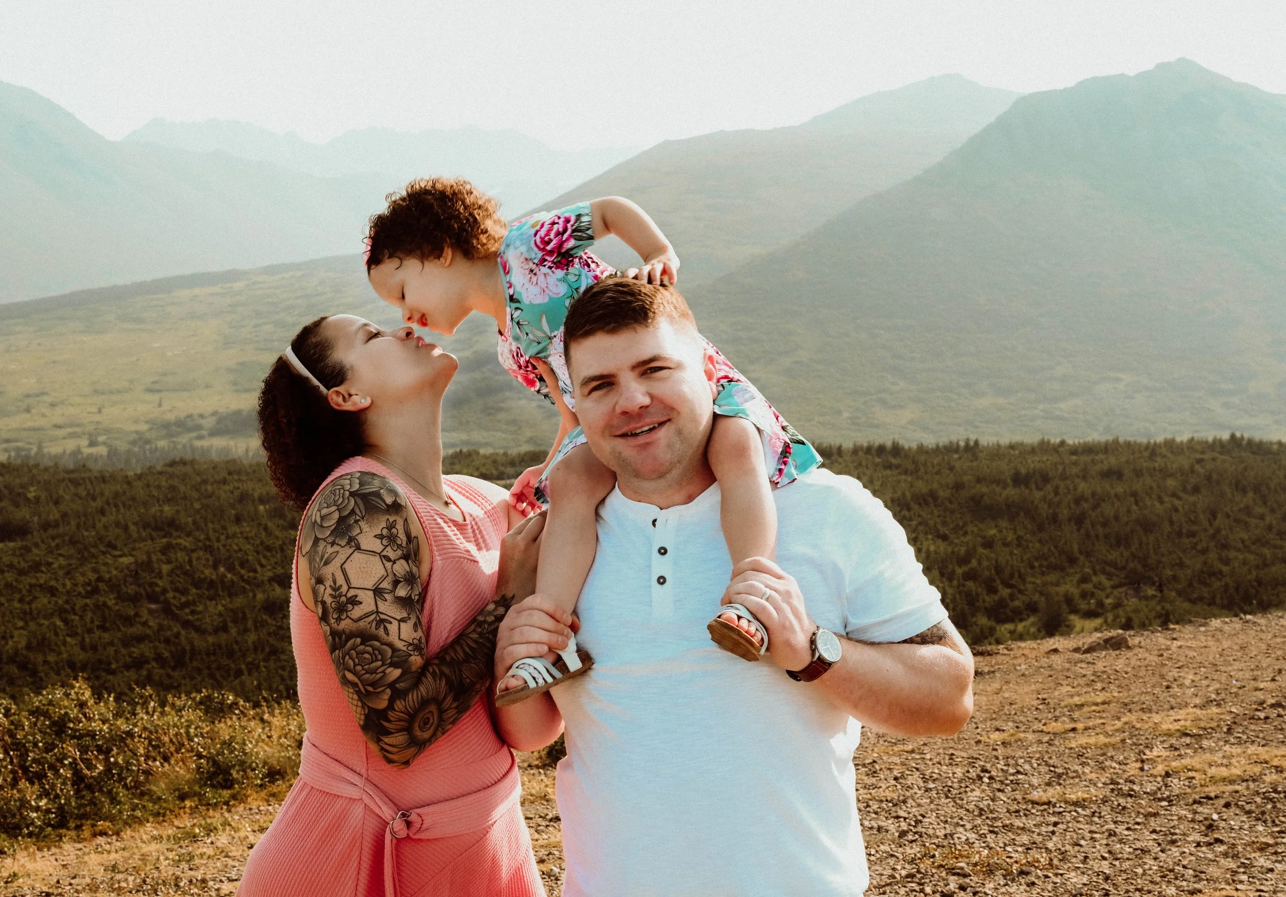 A family of three enjoying outdoors near mountains: a woman with tattoos on her arm and a man are holding a young girl on their shoulders; they are smiling and the girl is leaning toward the woman, about to kiss her.
