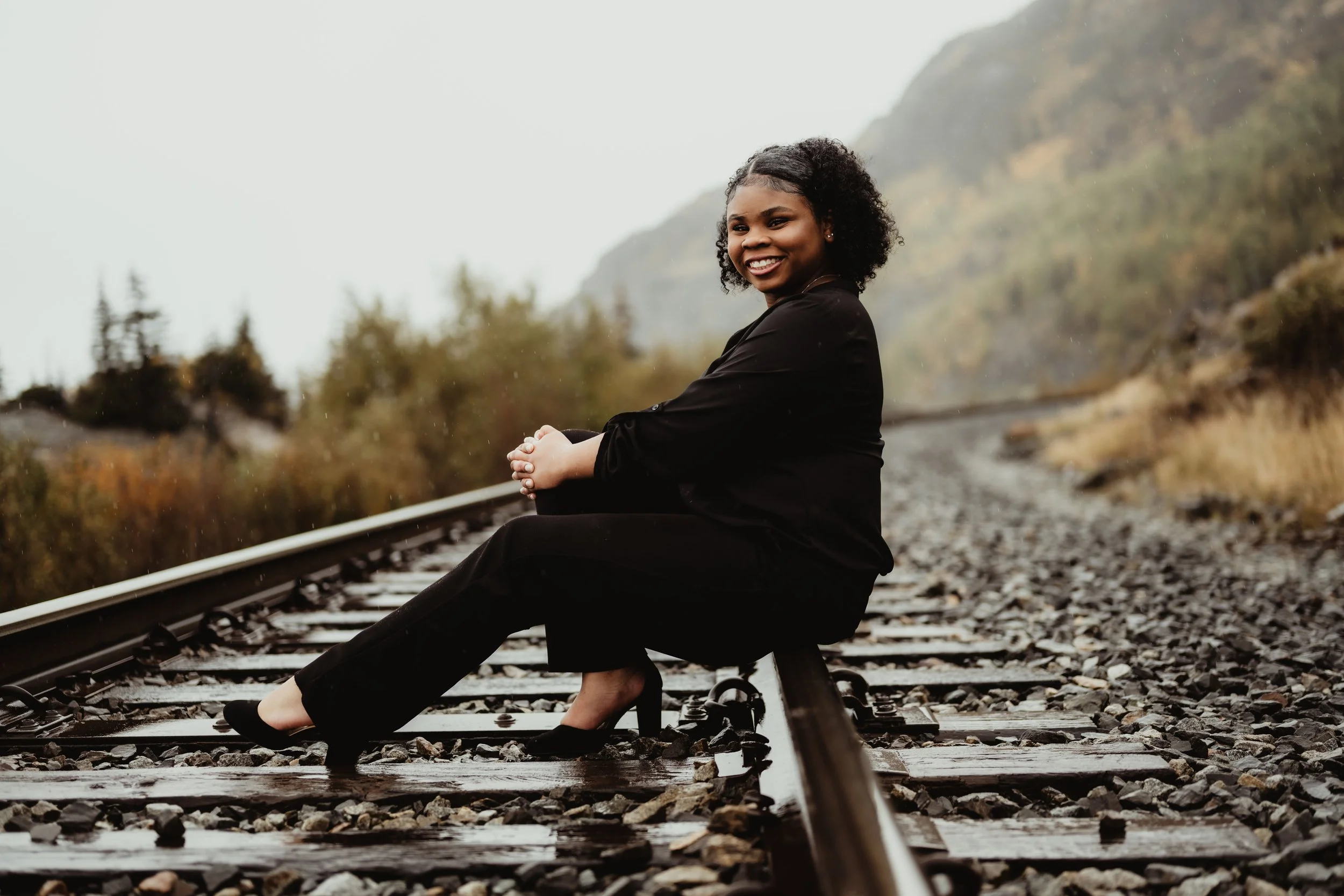 A woman sitting on railway tracks outdoors during rainy weather, smiling and looking at the camera, with a background of trees and mountains.