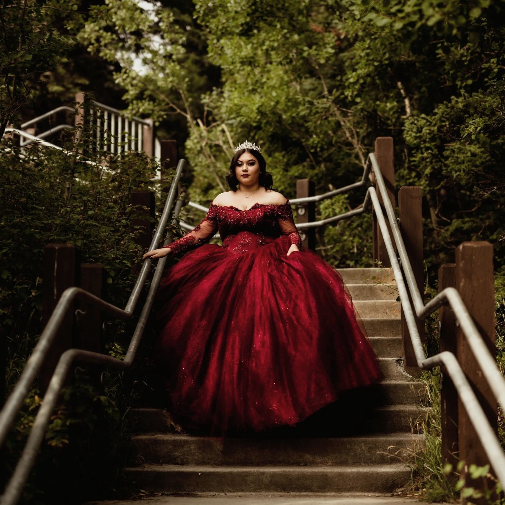 A woman in a red ball gown with an off-shoulder neckline and long lace sleeves, sitting on outdoor stairs surrounded by greenery, wearing a tiara and makeup.