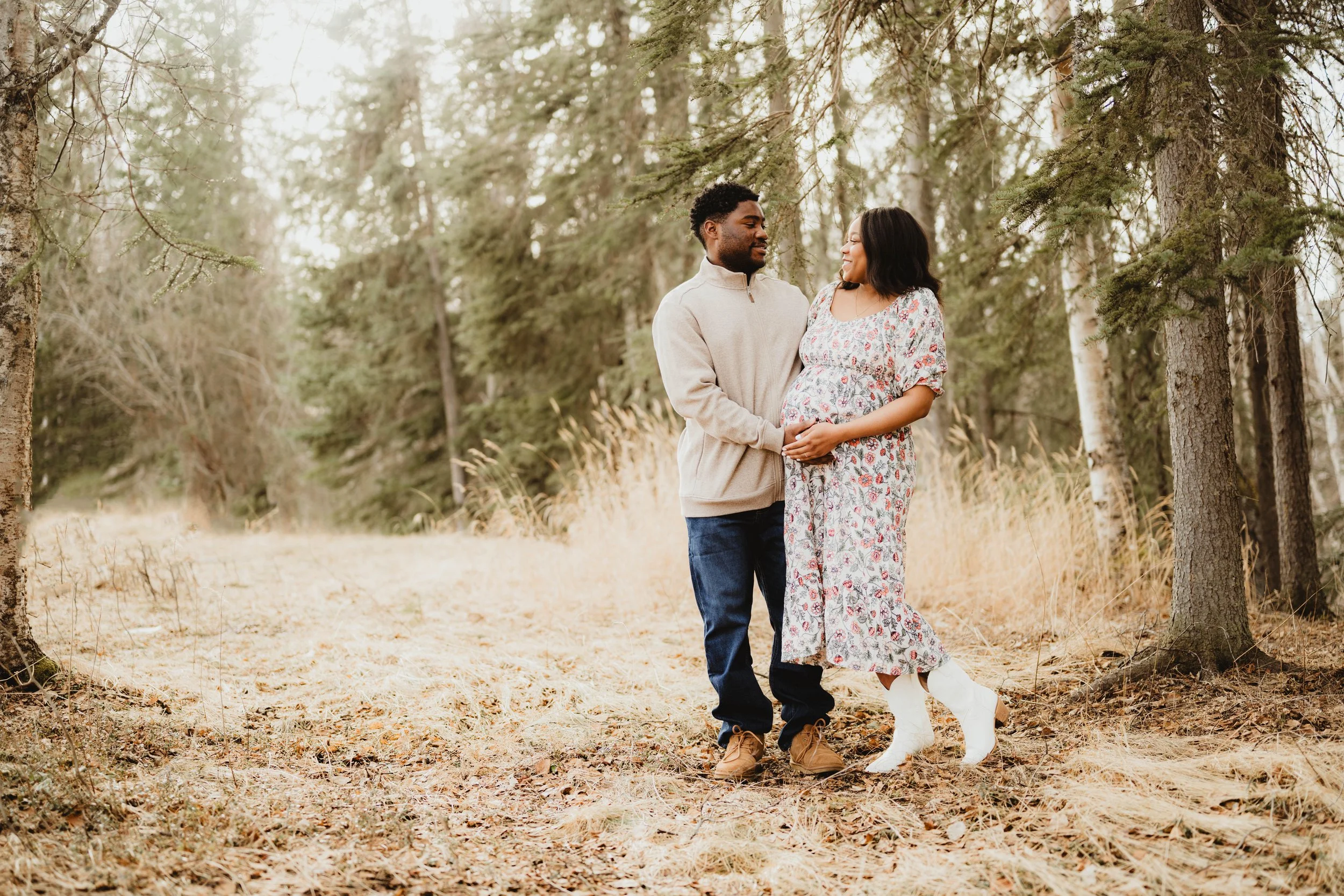 A pregnant woman in a floral dress and white boots standing on a forest trail, holding her belly, with a man in a beige sweater and jeans standing beside her, both smiling and looking at each other.