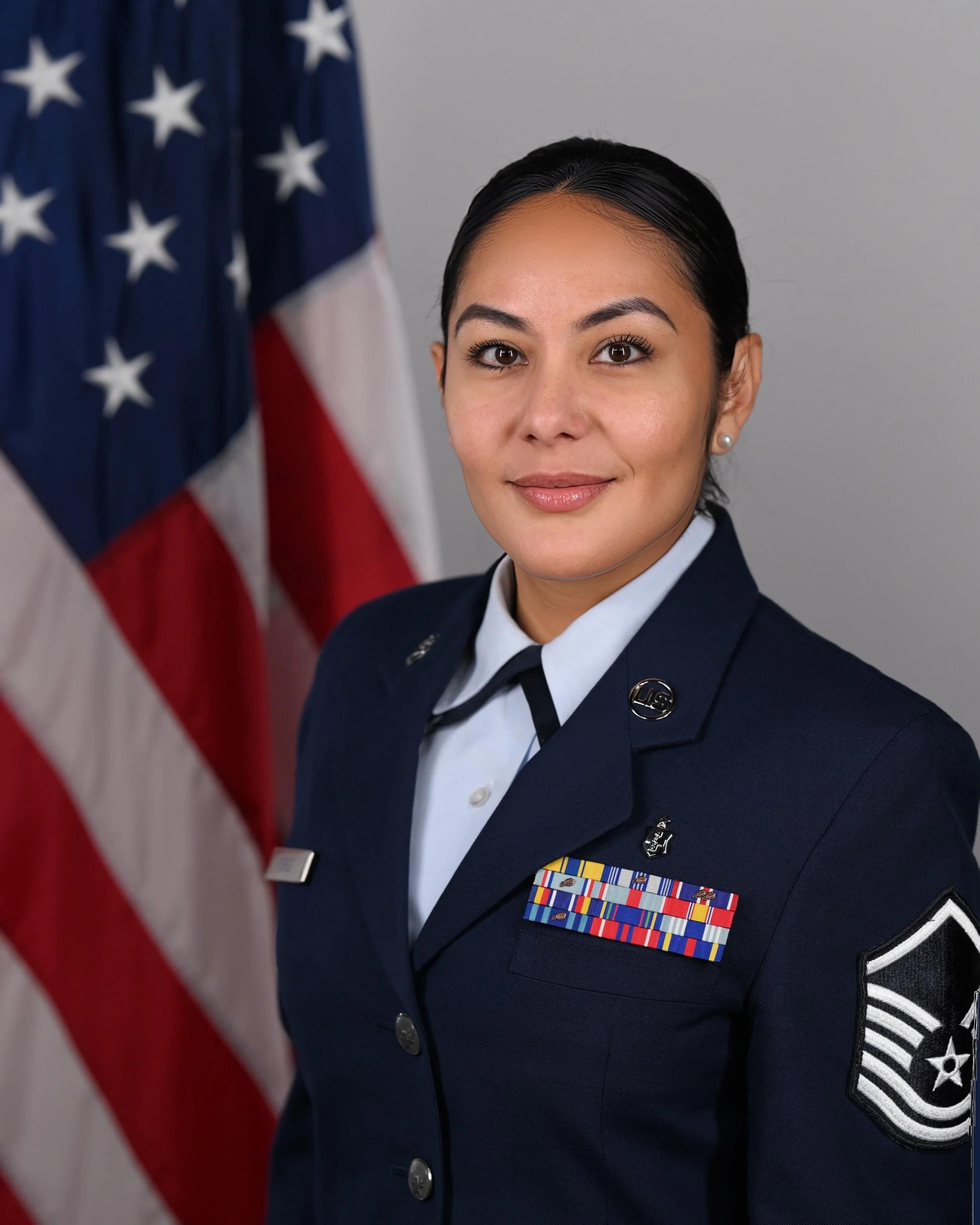 A female soldier in uniform standing in front of American flags, looking at the camera.