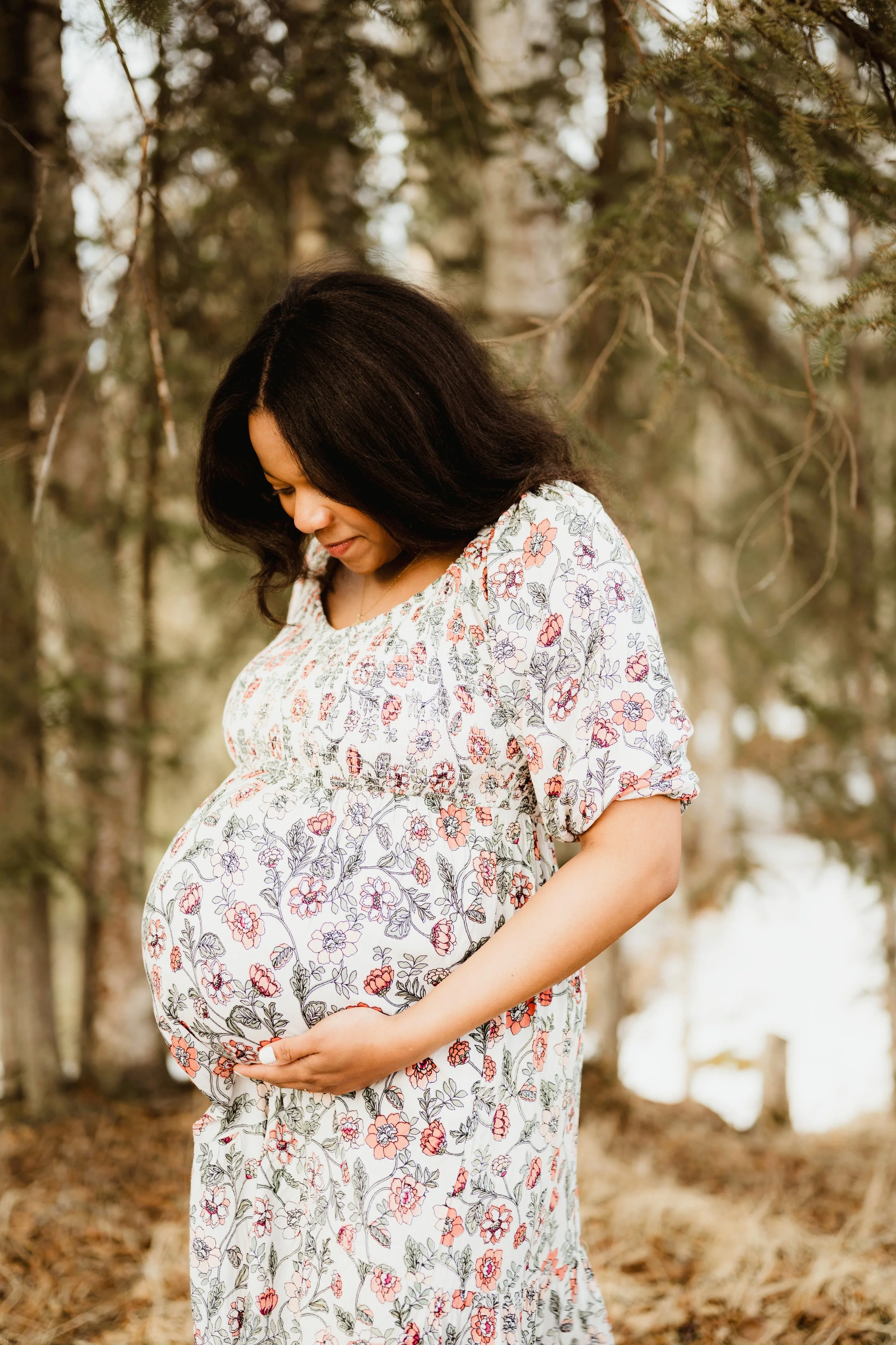 A pregnant woman in a floral dress standing outdoors among trees, gently holding her baby bump and looking down.