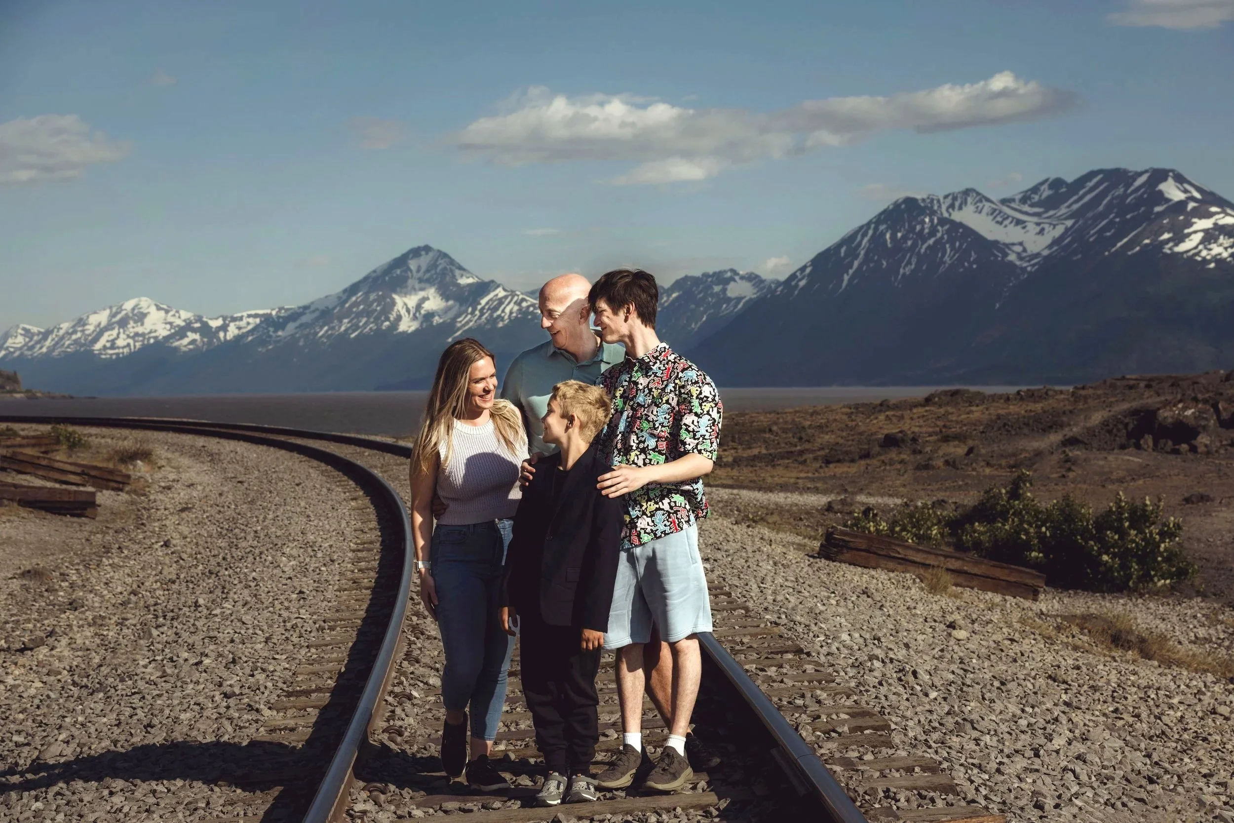 A family of five standing on railroad tracks in a mountainous landscape with snow-capped peaks and a lake in the background, enjoying a sunny day.
