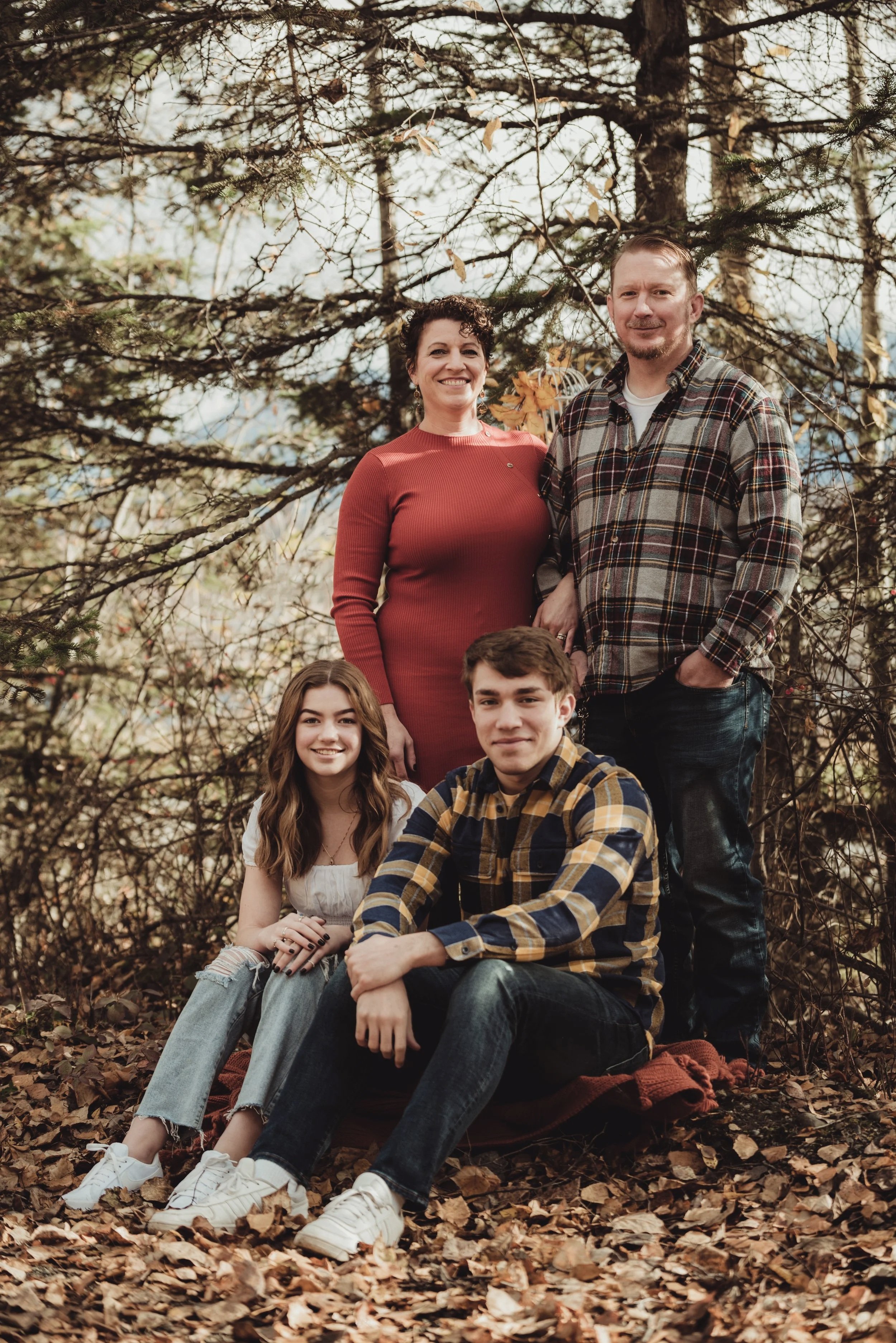 A family of four outdoors in a wooded area during fall, with leaves on the ground and trees in the background. The mother wears a red dress, the father a plaid shirt, the daughter a white top and ripped jeans, and the son a plaid shirt and dark jeans. They are smiling and posing for the photo.