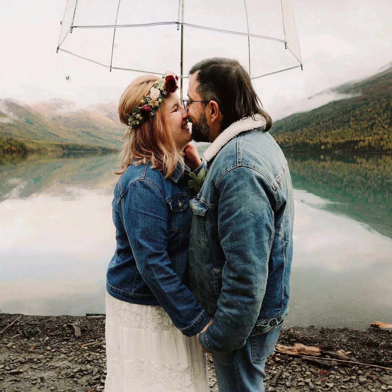 A couple in denim jackets sharing a kiss under a transparent umbrella near a lake and mountains.