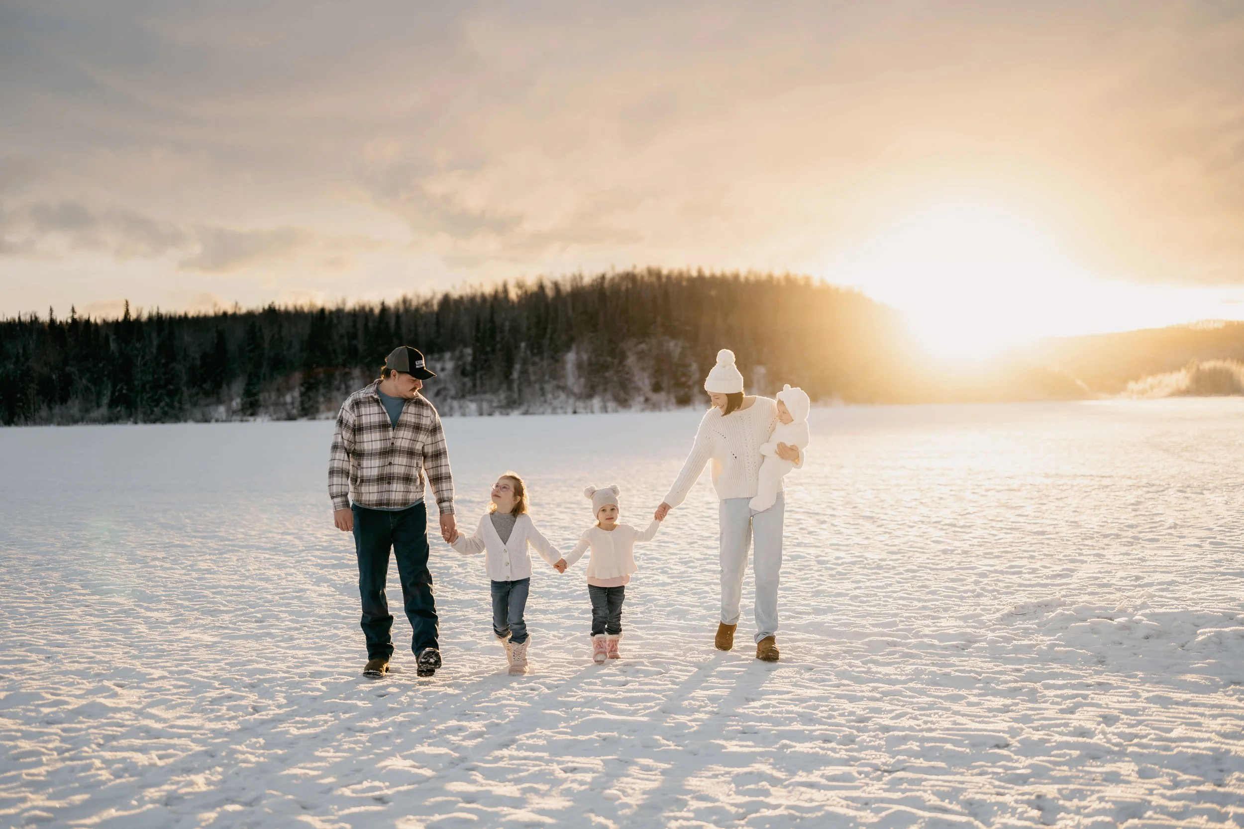 Family of four walking hand-in-hand on snowy landscape during sunset, with a forest in the background.