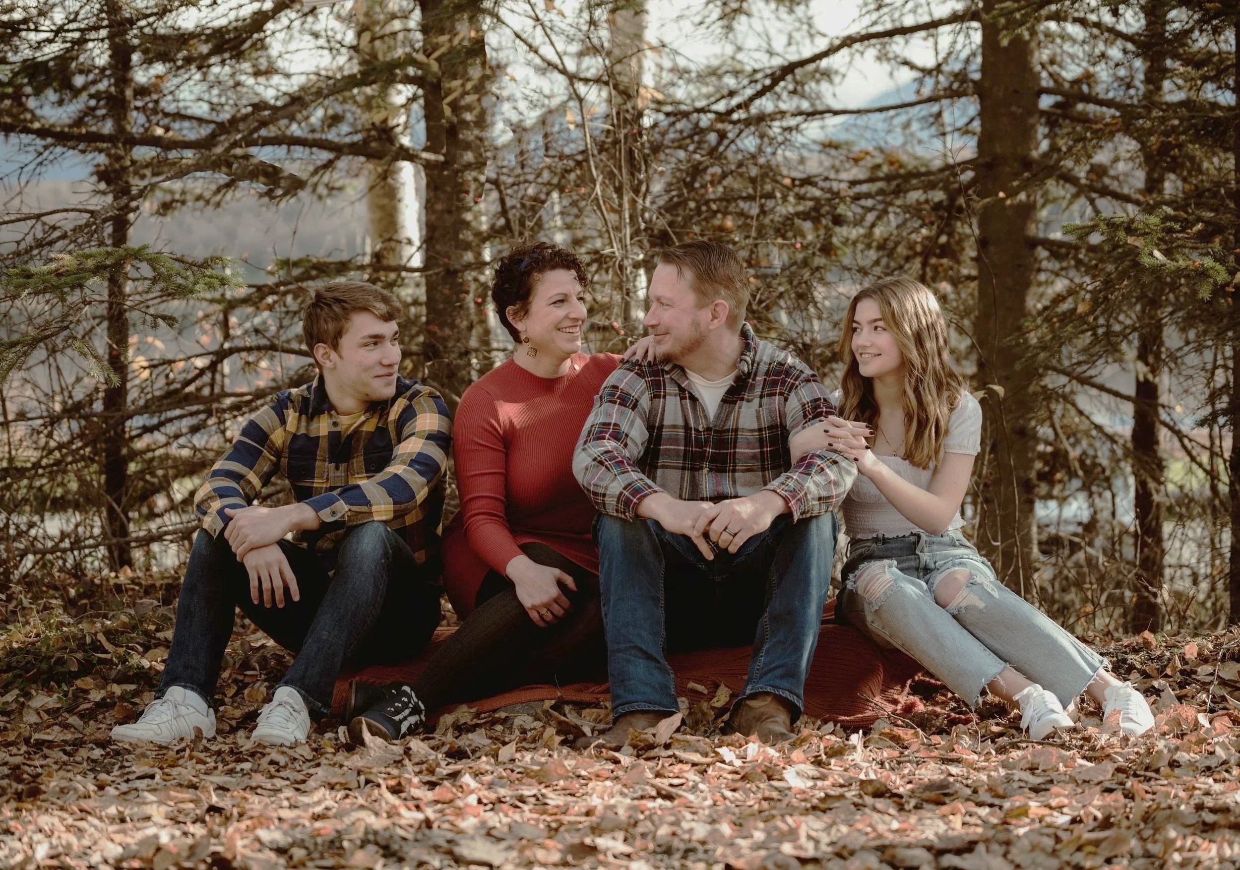 A family of four sitting on a blanket in a wooded area with autumn leaves. They are smiling and engaging with each other.