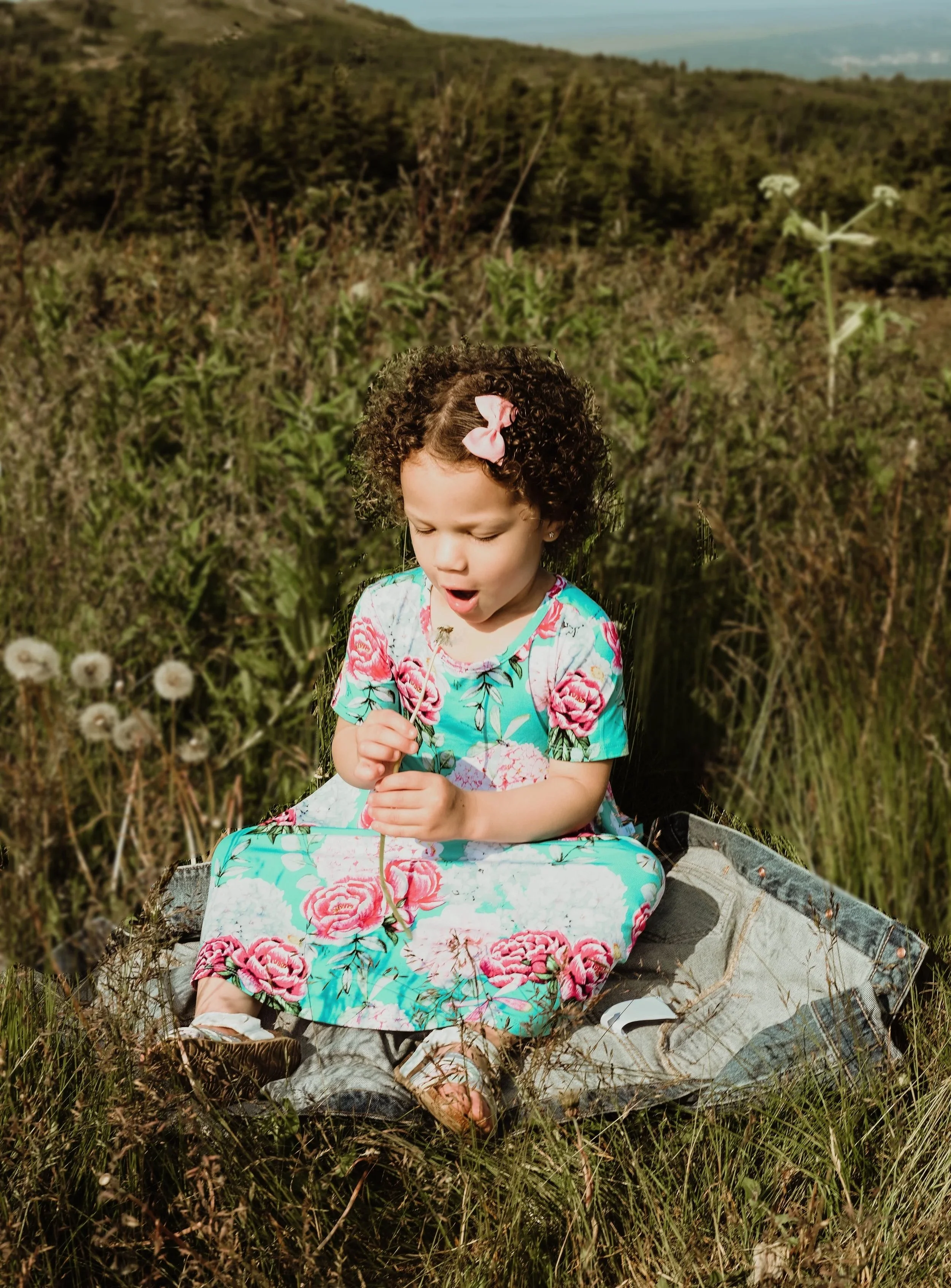 A young girl with curly hair and a pink bow, wearing a floral dress, sits on a denim jacket in a grassy field, holding a dandelion and blowing its seeds.