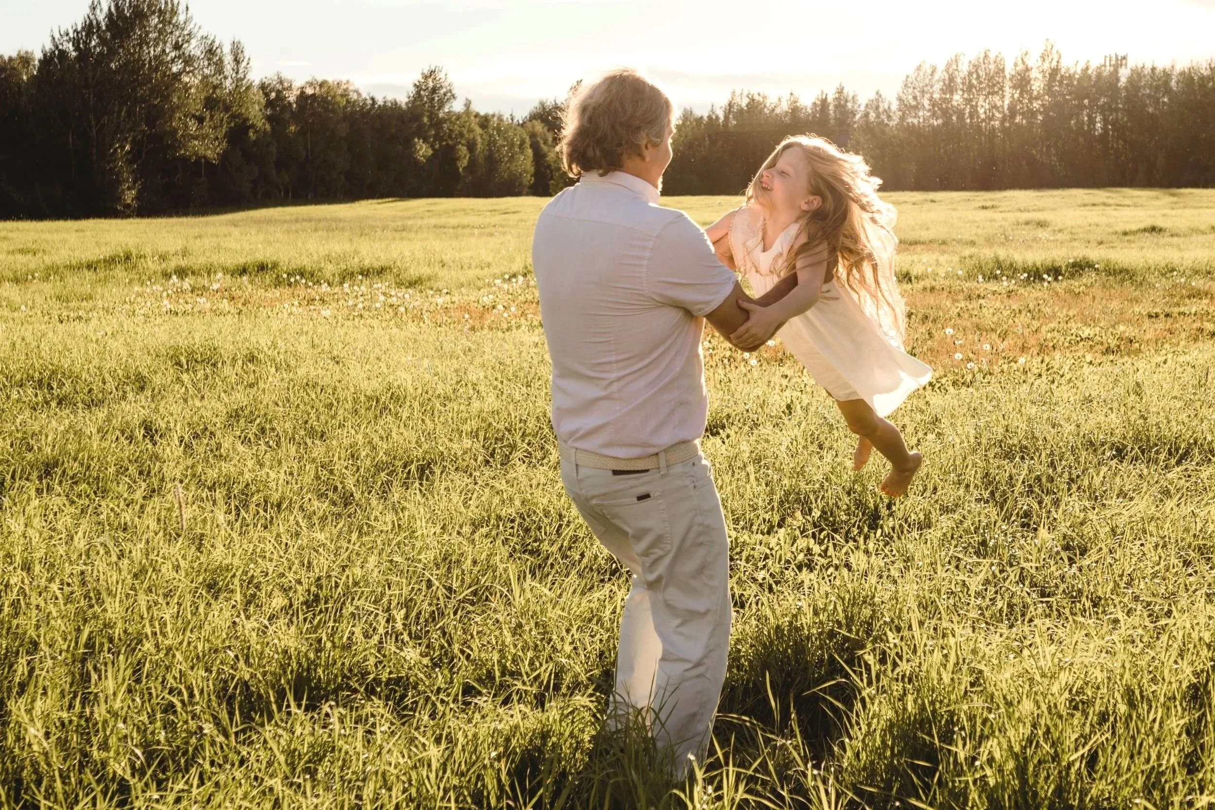 A man lifting a young girl in a grassy field during sunset, smiling and playing.