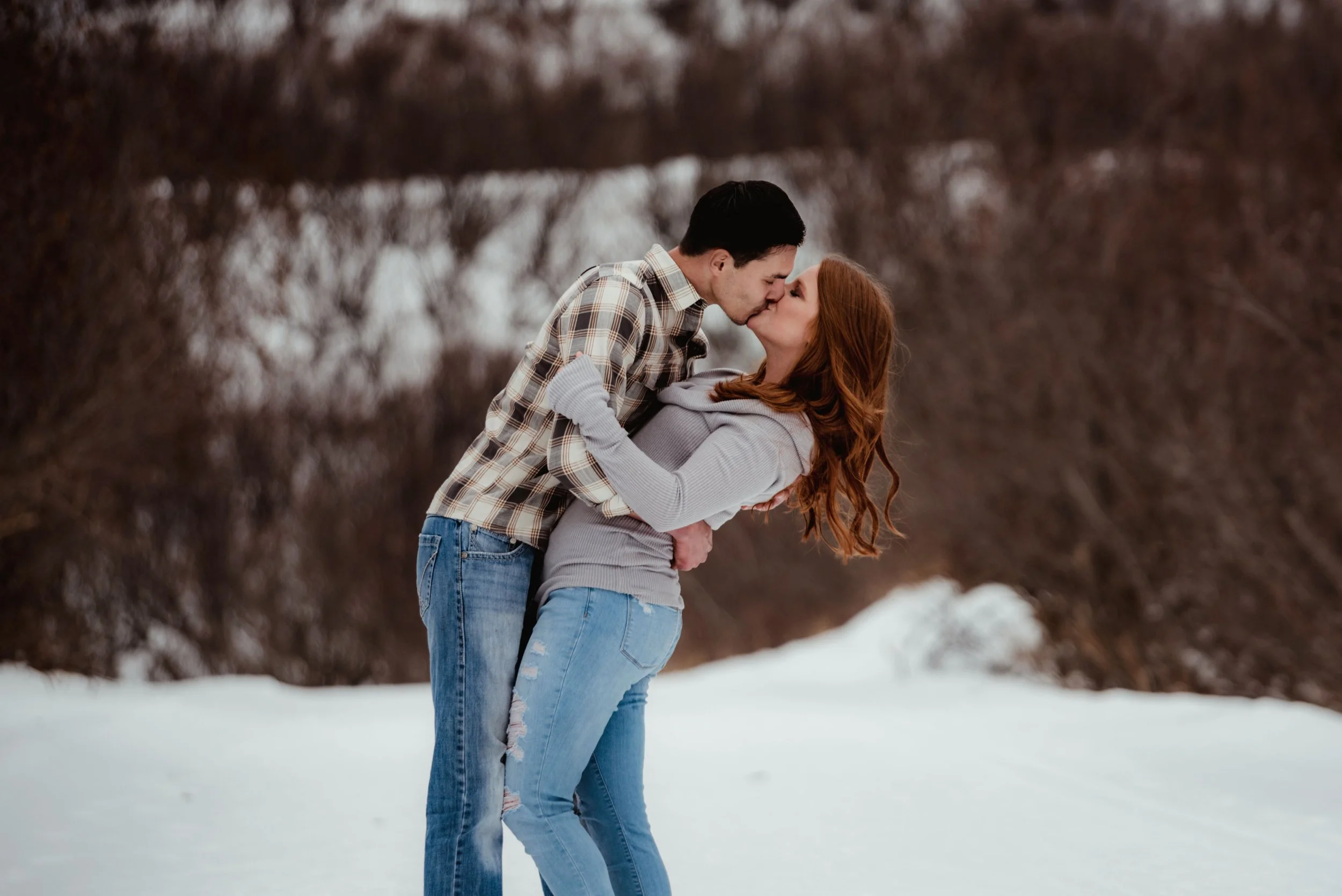 A couple kissing outdoors in a snowy landscape during winter, with the man leaning over the woman who has long red hair, both wearing casual winter clothing.