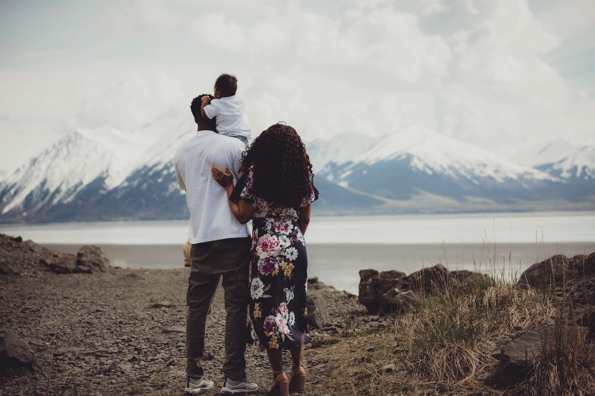 A family of three stands on rocky terrain near a lake with snow-capped mountains in the background. The man is holding a young child on his shoulders, and a woman stands beside them, facing away from the camera.