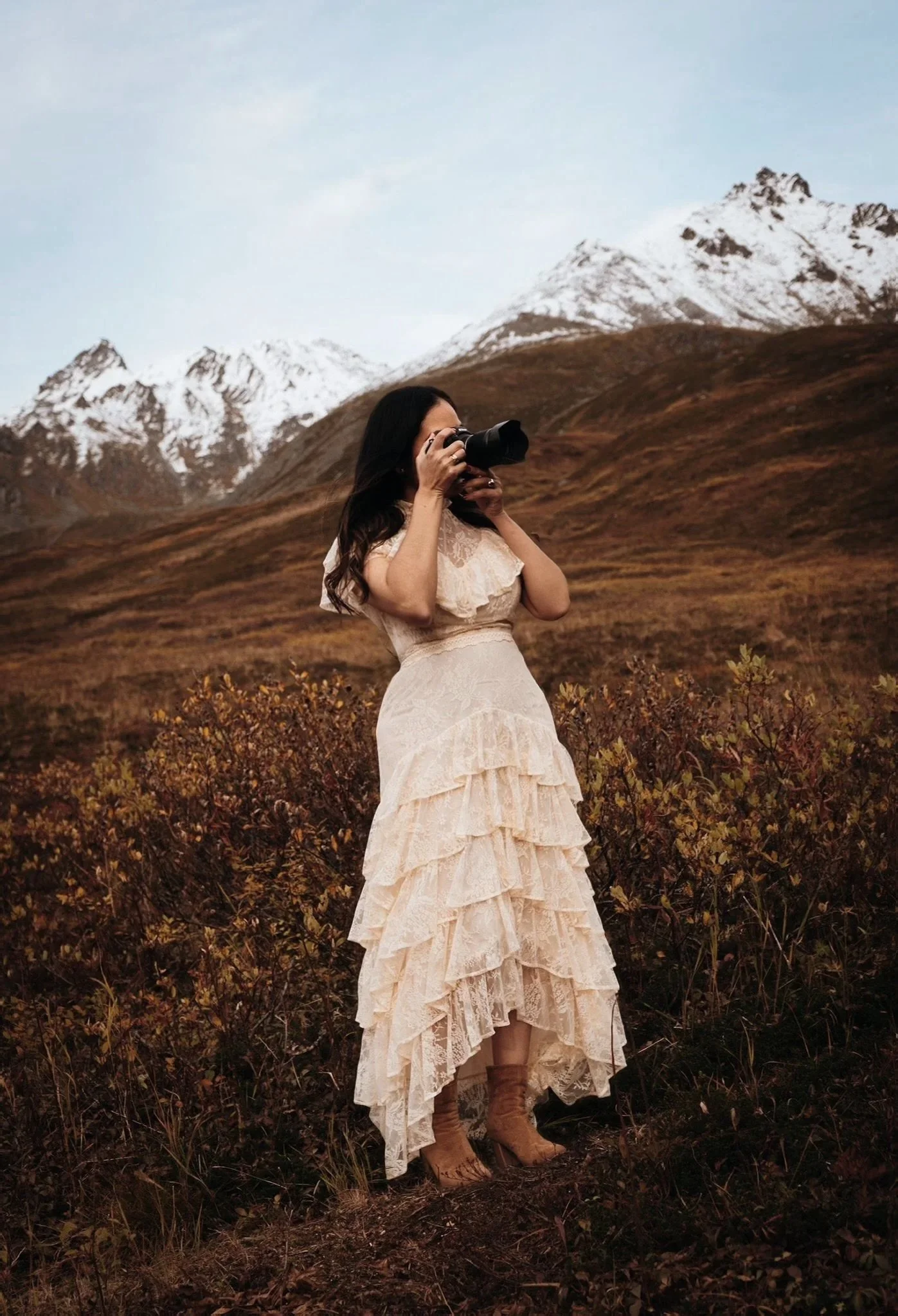 A woman in a long, ruffled, cream-colored dress and brown boots taking photos with a camera in a mountainous landscape with snow-capped peaks and brown rolling hills.