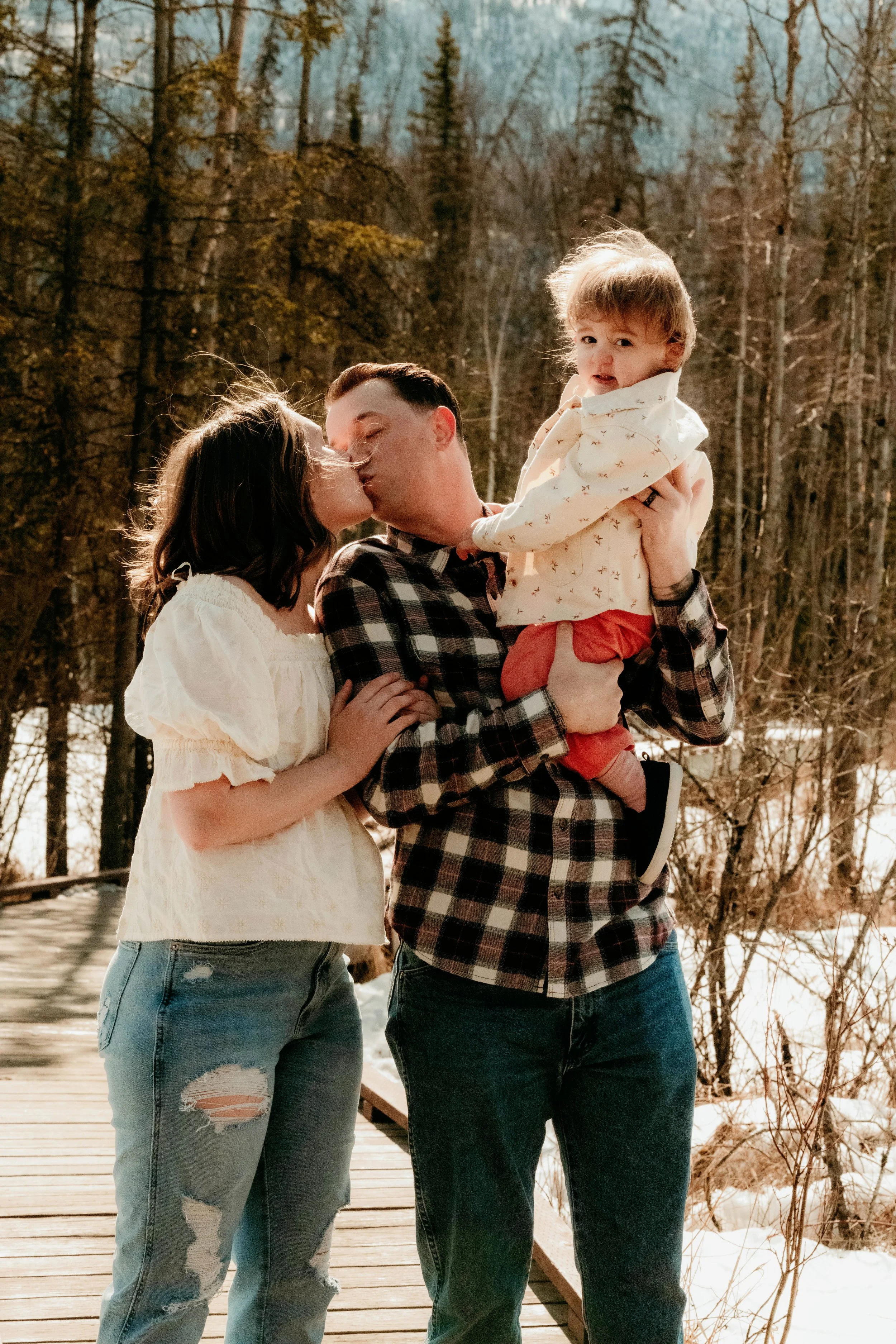 A family of three outdoors in a wooded area with snow, with the father holding a child and the mother kissing the father.