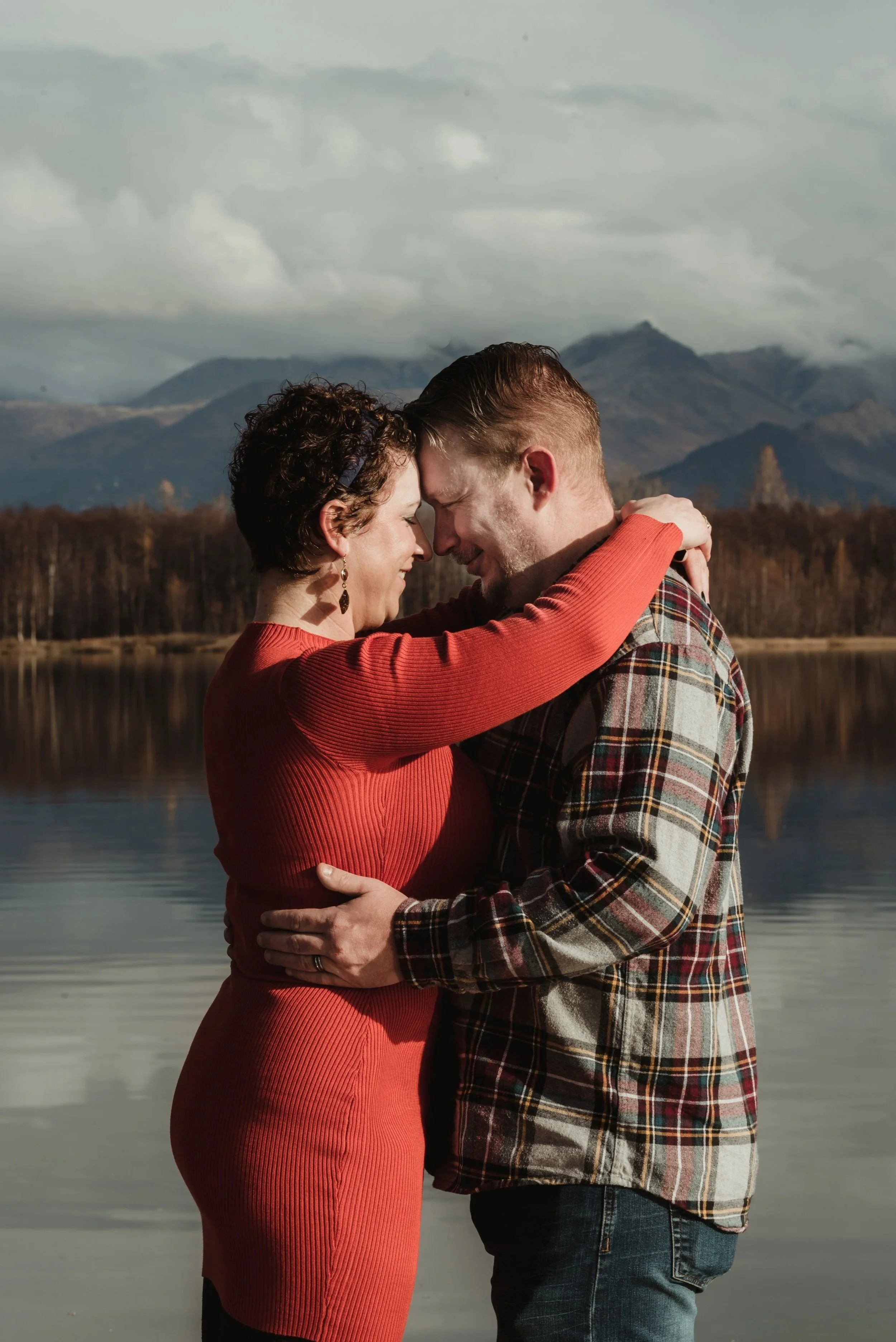 A couple embracing each other by a lake with mountains in the background.