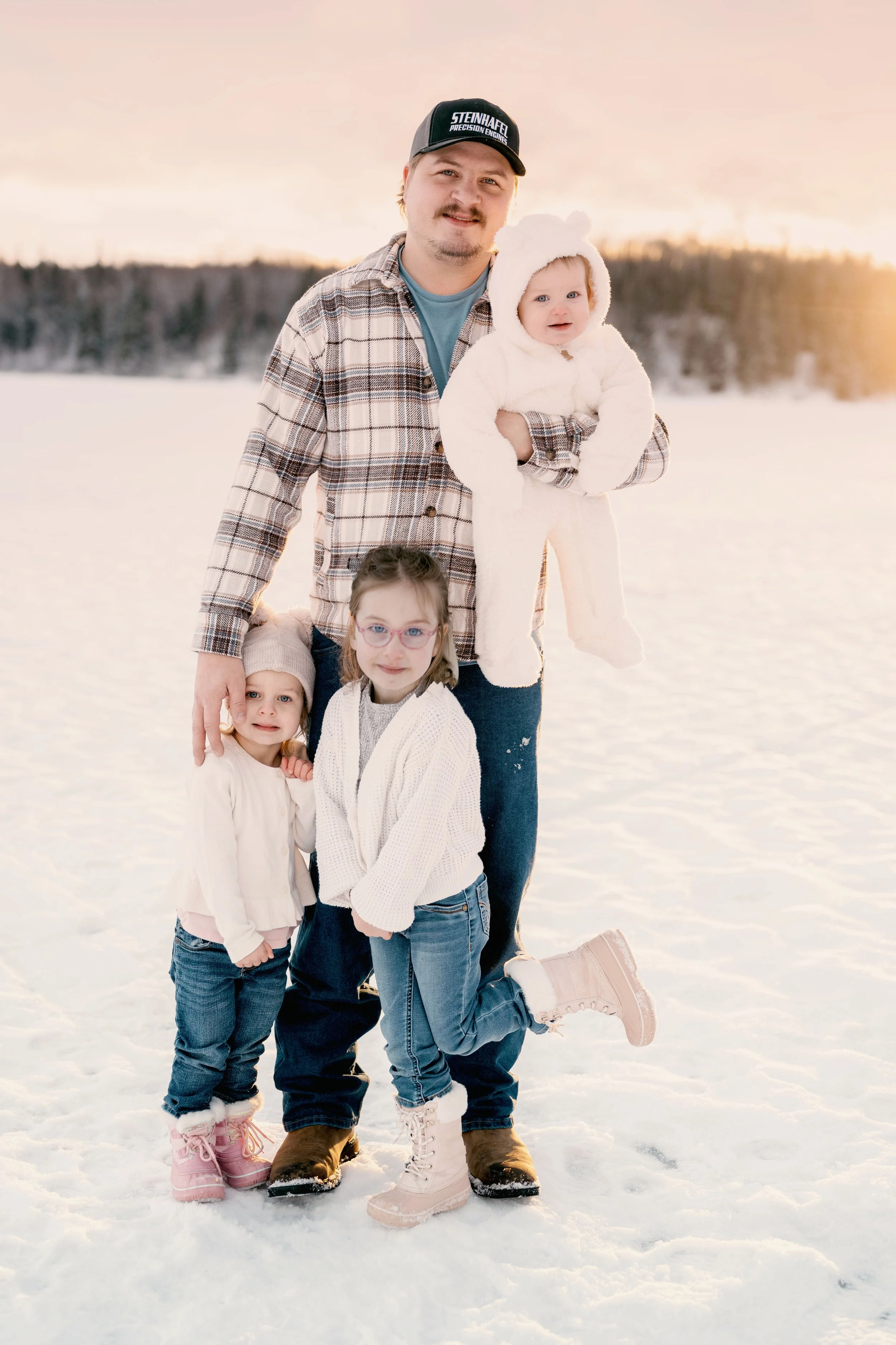 A man and three young girls standing on snow-covered ground outdoors during sunset. The man is holding a baby girl dressed in a white fuzzy outfit with bear ears. The other two girls, one with glasses and the other wearing a hat, stand at his feet. The background features a forest and a warm, orange-pink sky.