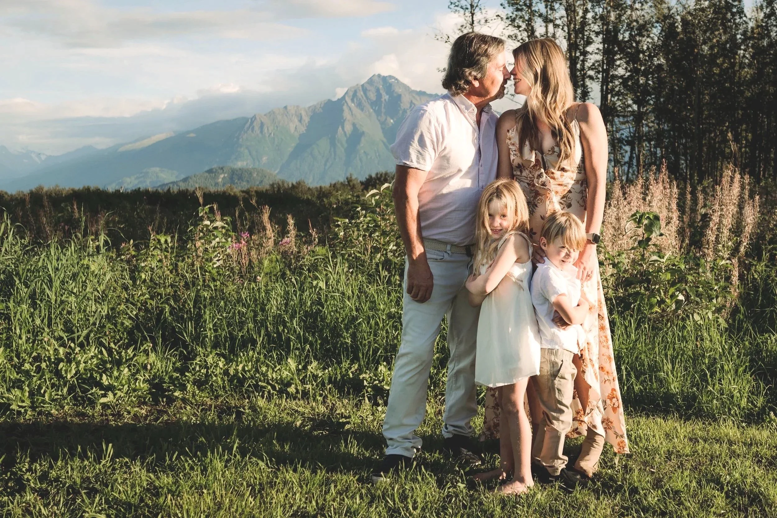 Family of five, including two adults and three children, standing outdoors on a grassy field with mountains and trees in the background, enjoying sunlight, during a scenic outdoor setting.